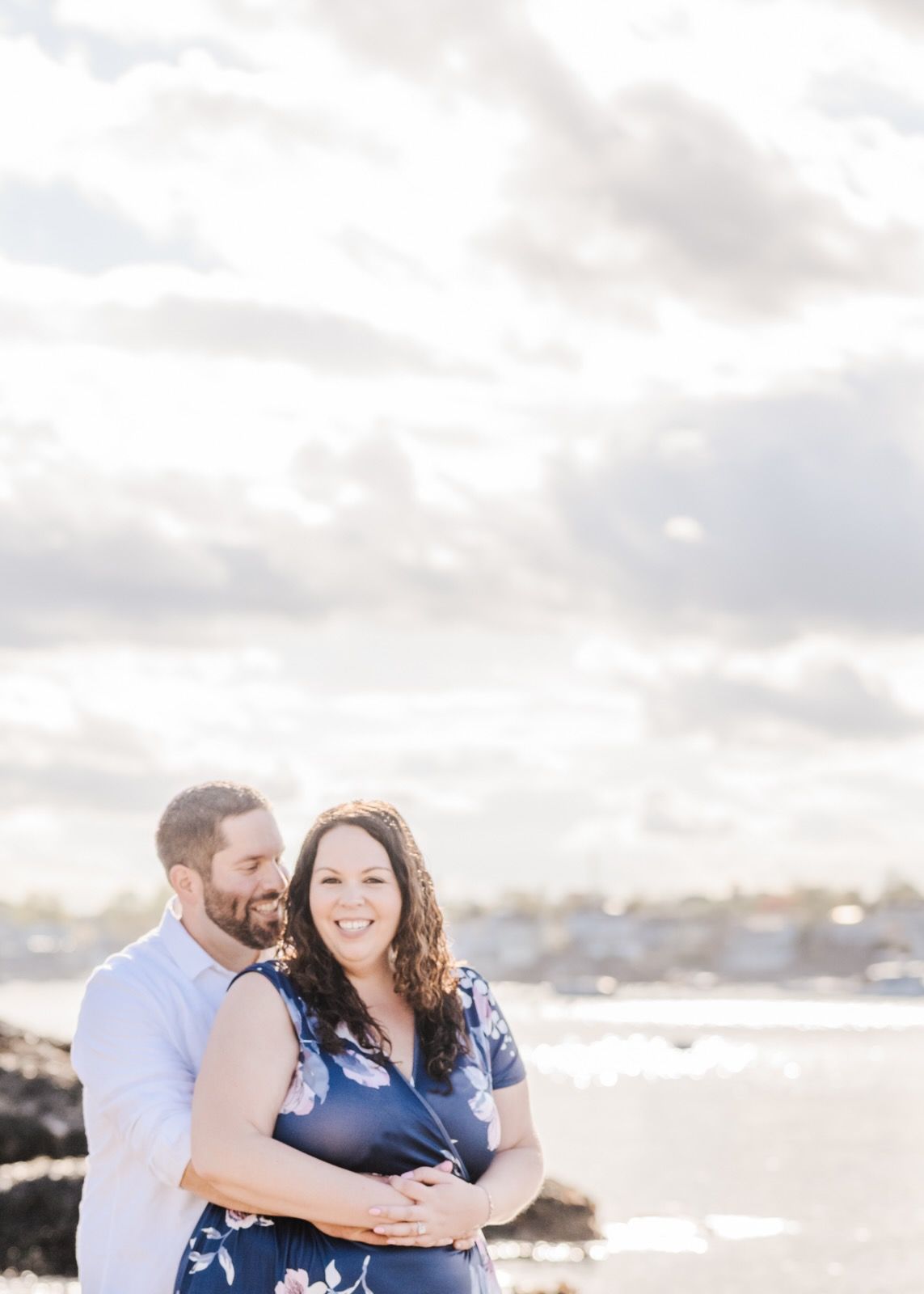 A couple poses together on a rocky beach with city skyline visible in the distance.