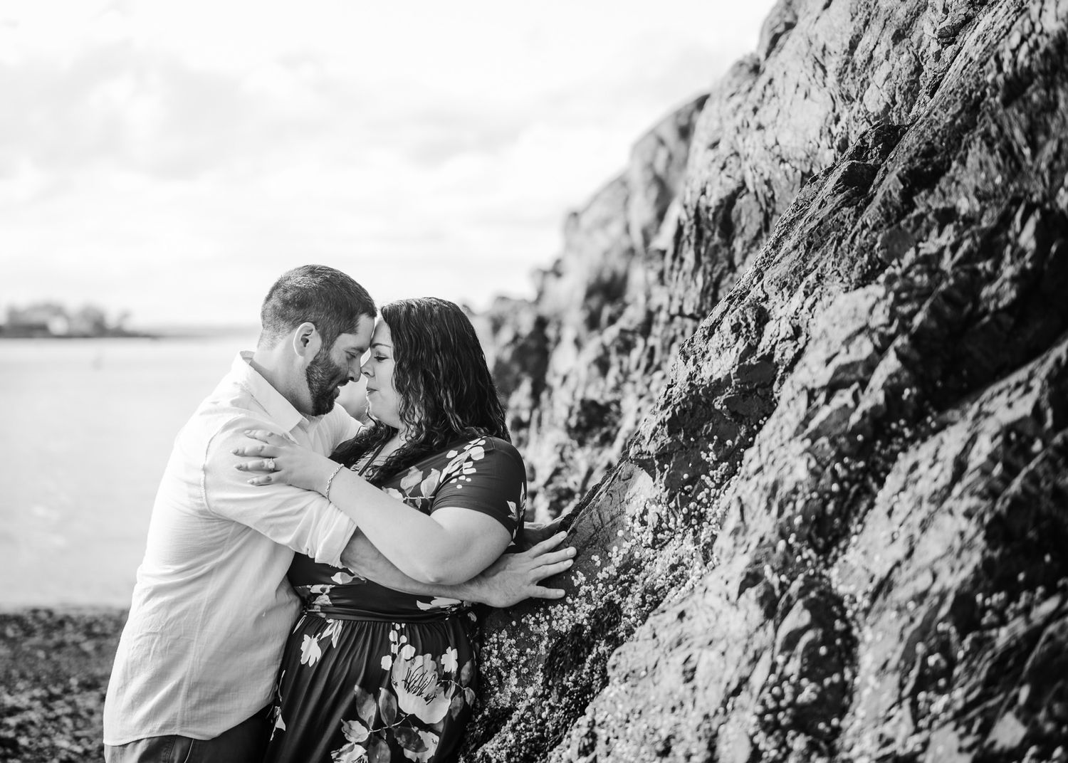 A romantic black and white portrait of a couple embracing against rocky cliffs by the ocean.