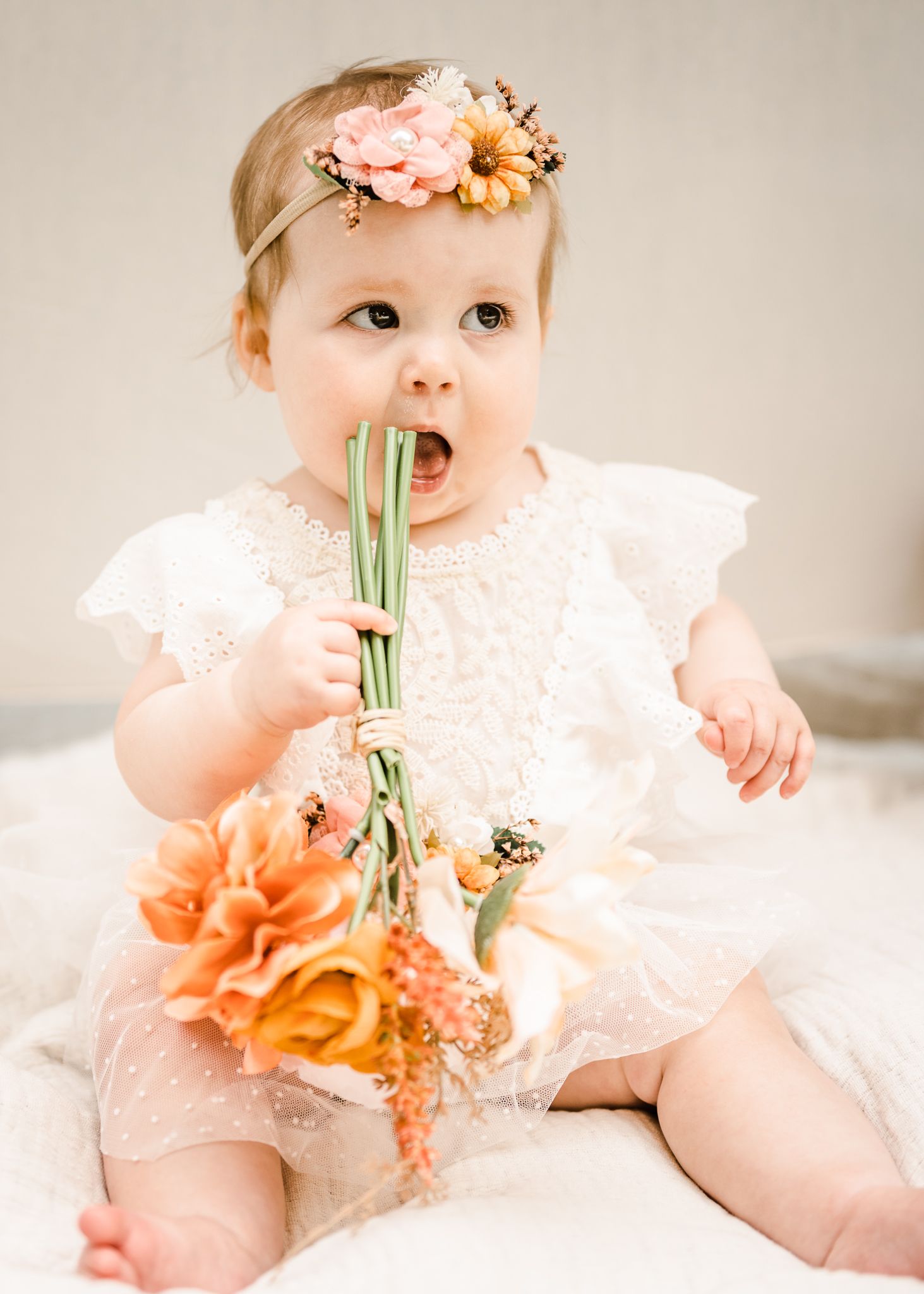 Baby in white lace dress holds bouquet of flowers while sitting on white fabric.
