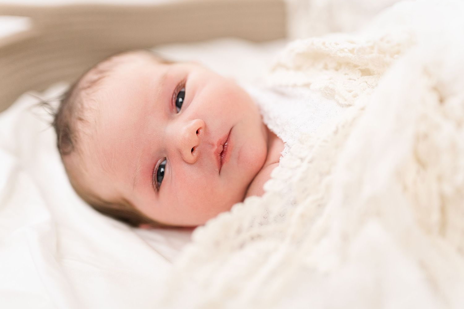 Sweet baby wrapped in soft white blanket looking directly at camera with bright eyes.