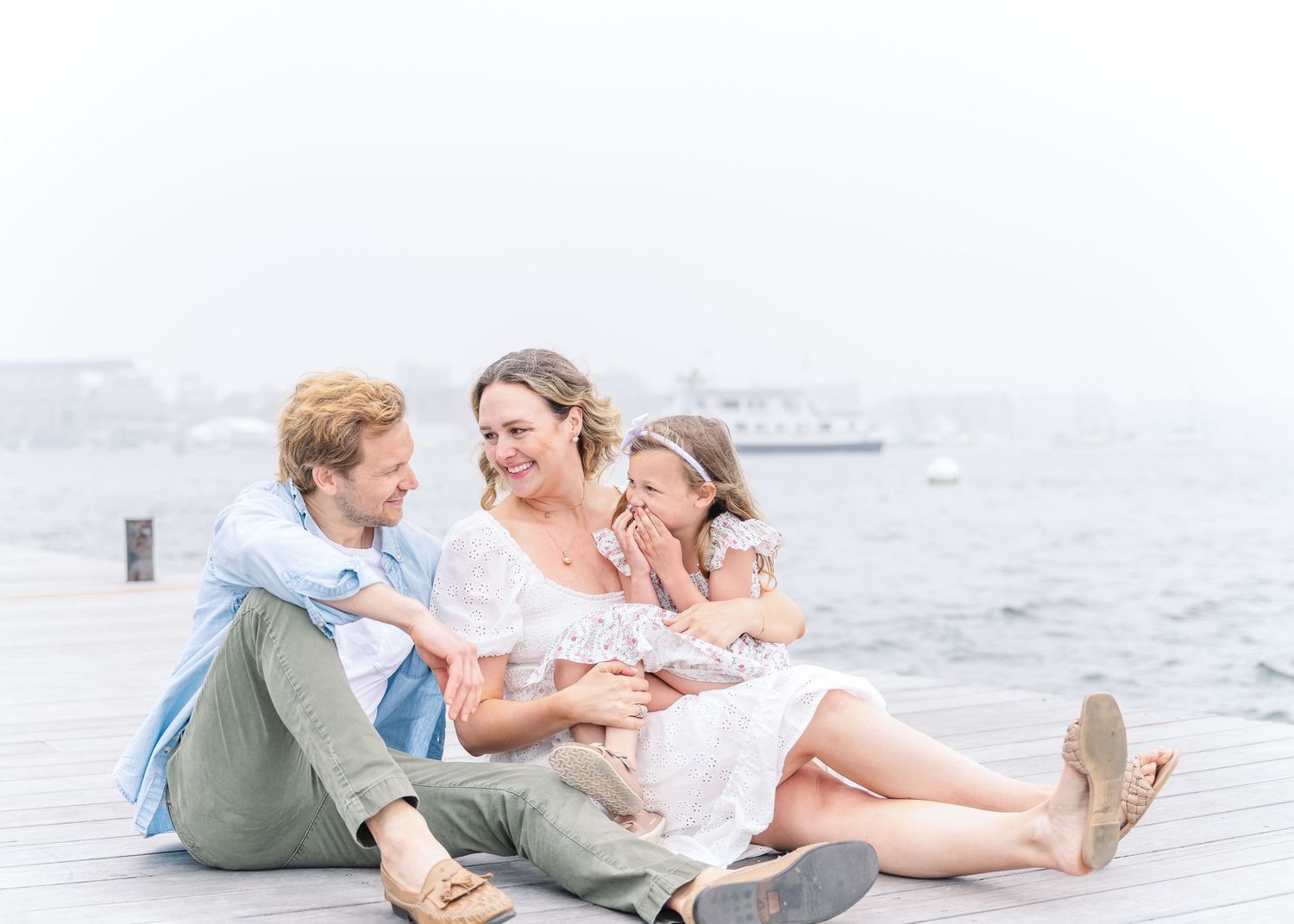 A relaxed family moment captured on a wooden dock by the ocean on a bright overcast day.