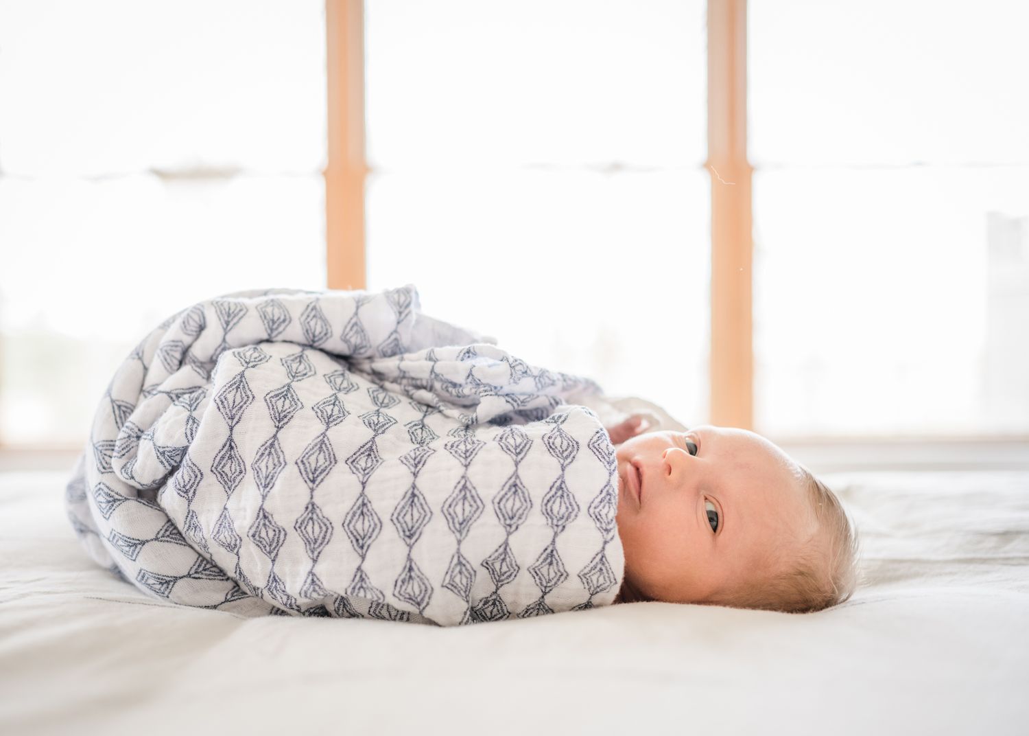 Baby wrapped in gray patterned swaddle lying on white bedding.