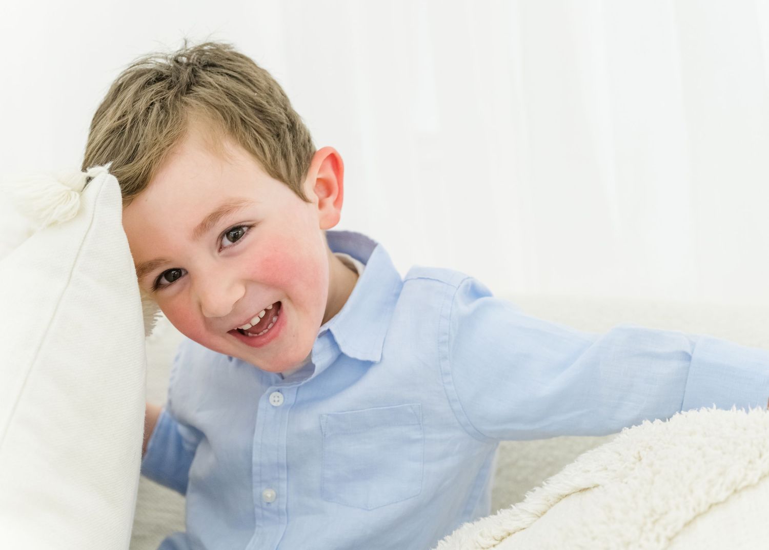 A cheerful child in a light blue button-down shirt lays on white cushions while smiling at the camera.