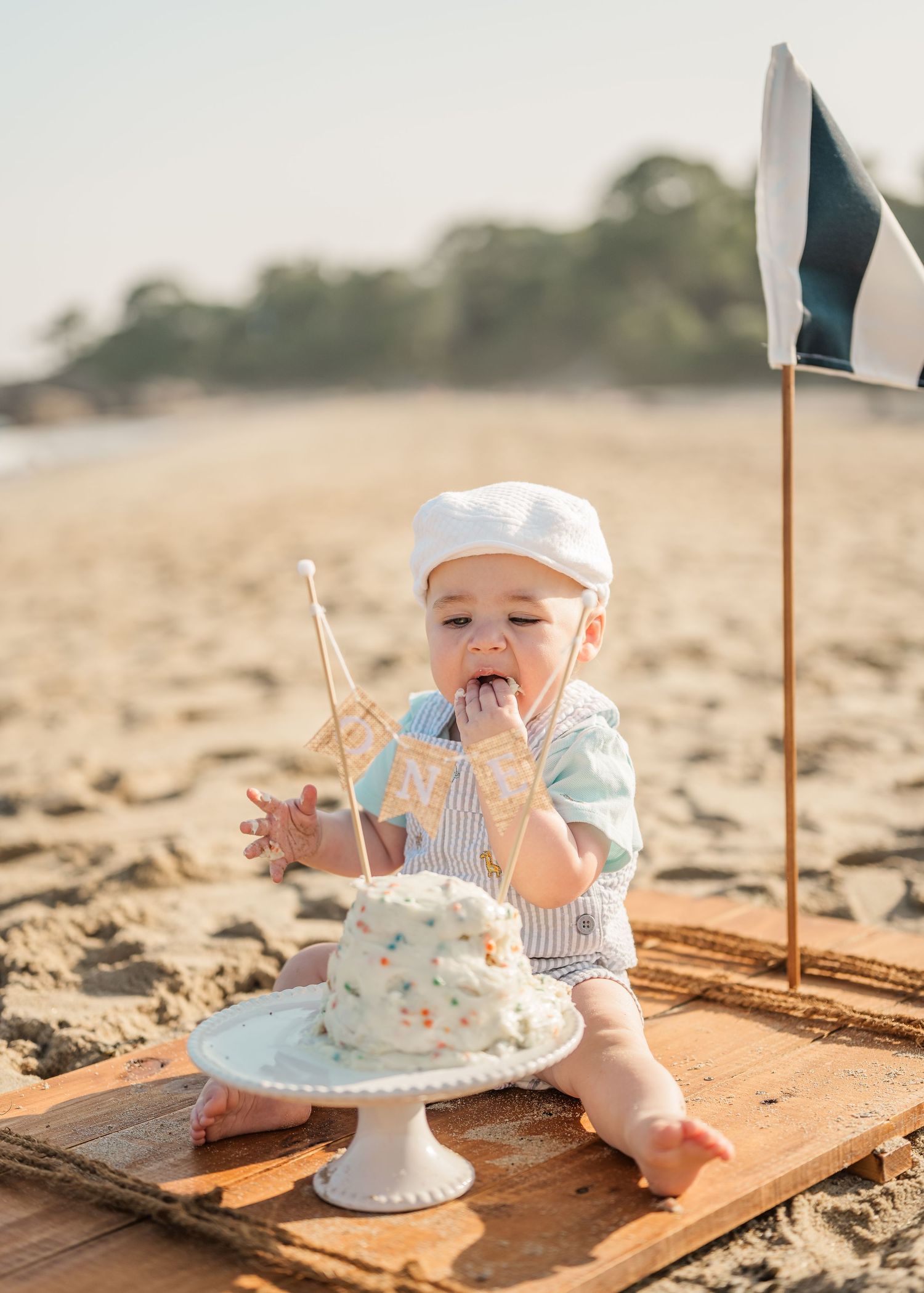 Baby in white hat enjoying first birthday cake smash photo session on sandy beach with rustic wooden backdrop.