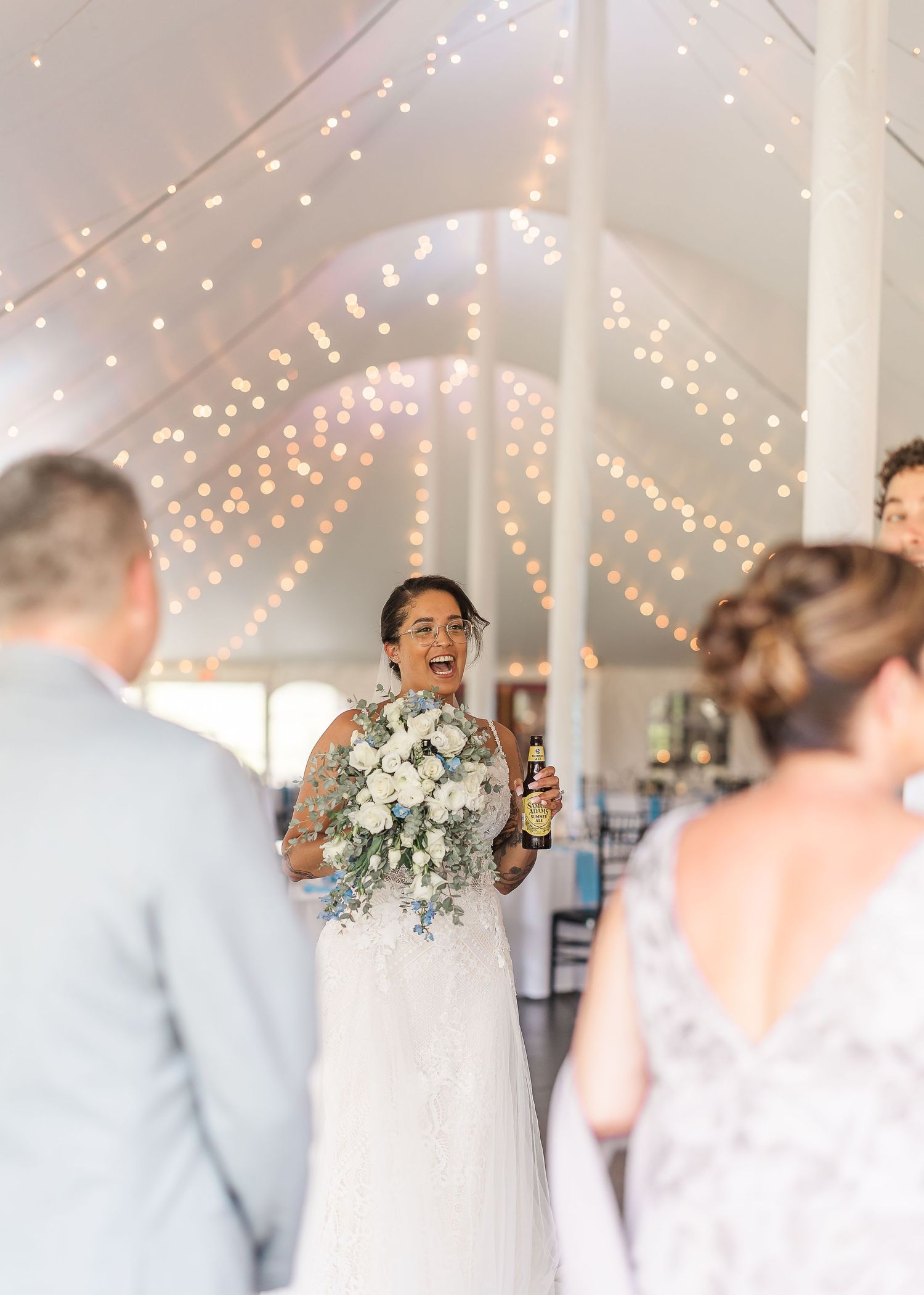 A bride in a white wedding dress dances and laughs joyfully under twinkling string lights at an indoor reception venue.