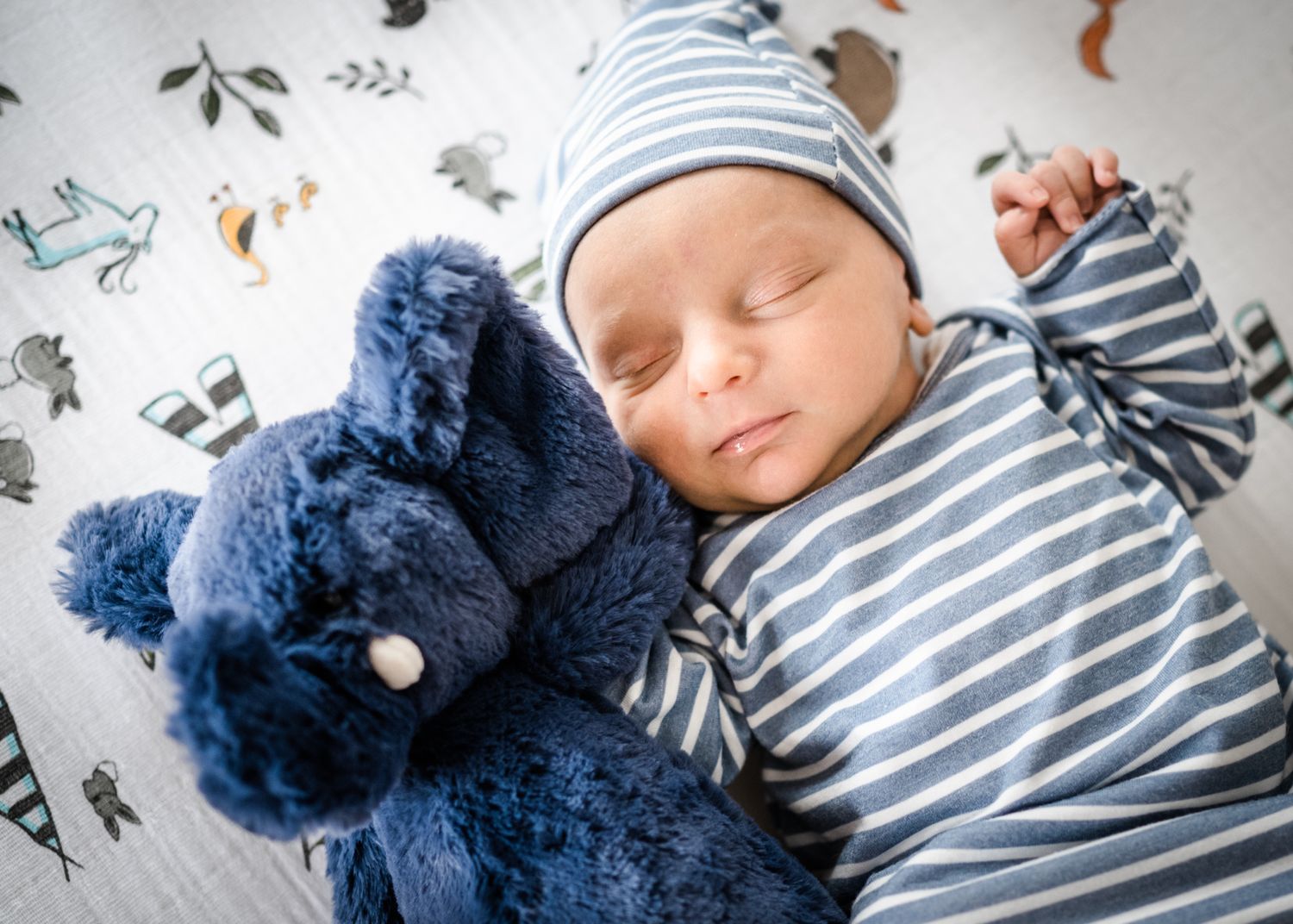 Sleeping baby in striped outfit cuddling with blue plush toy on patterned blanket.