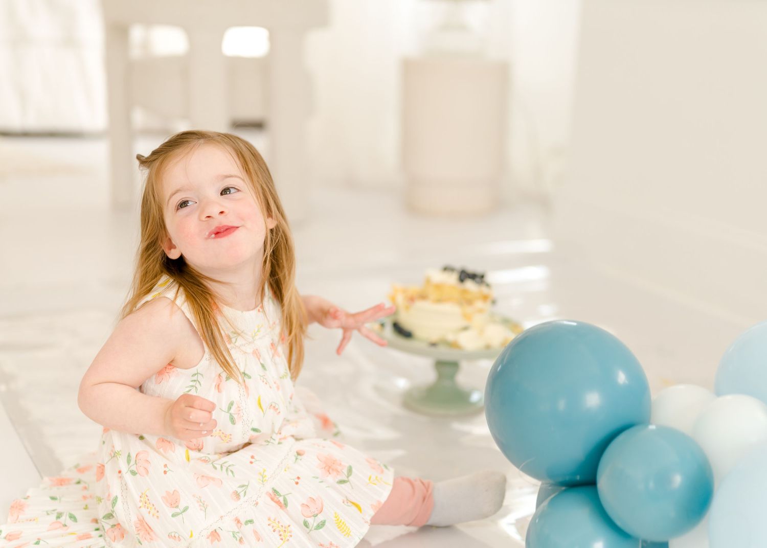 Young girl in a floral dress poses at a party setting with blue balloons and a cake stand.