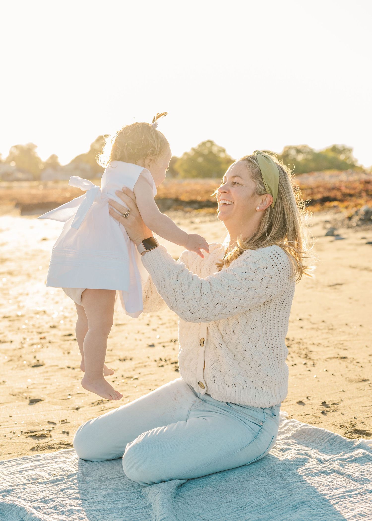 Parent and child share a playful moment on a blanket at the beach during sunset.