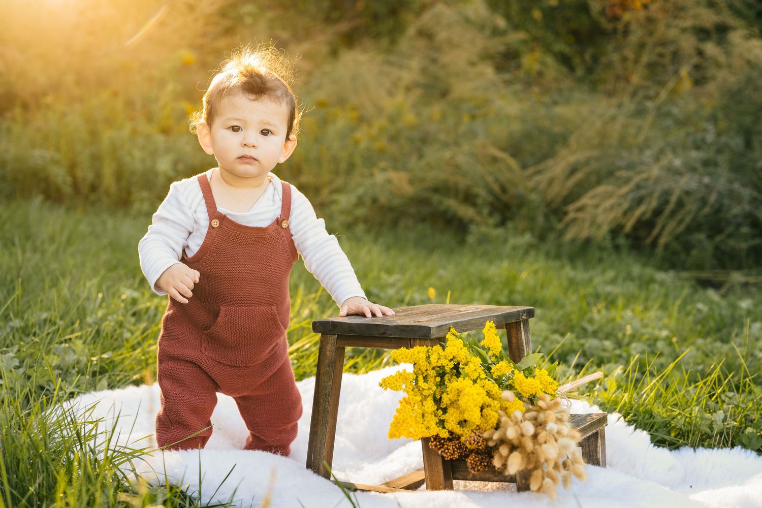 Outdoor photoshoot setup with rustic decorations and yellow flowers on a blanket in grass at sunset.