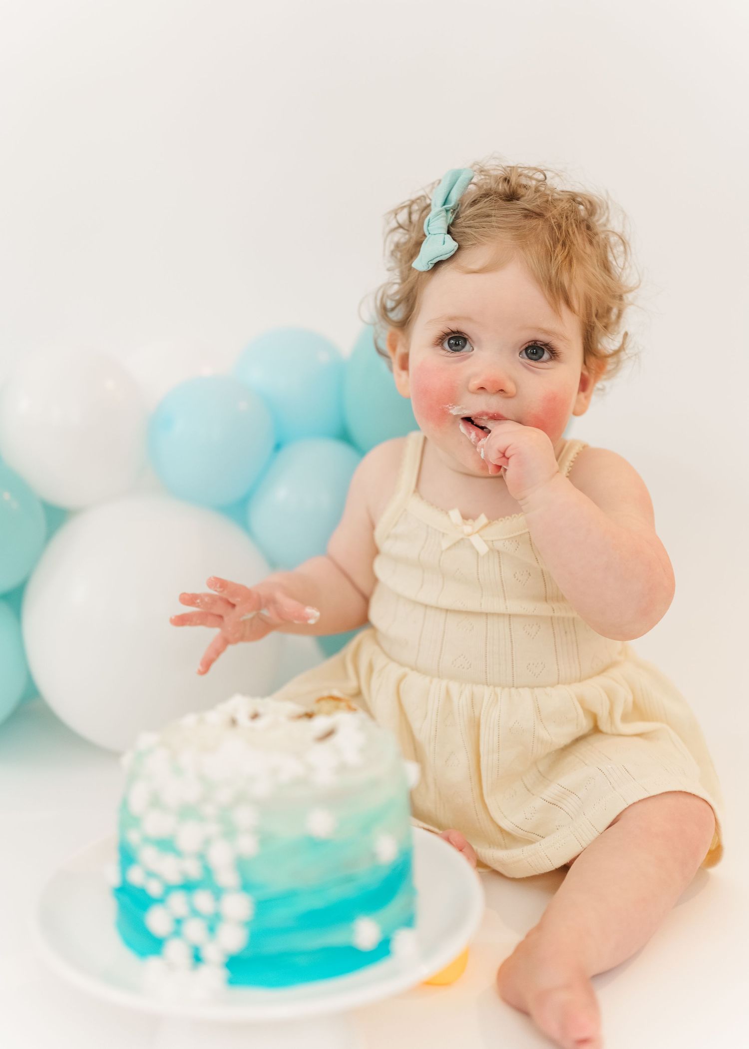 Cake smash. Baby in cream-colored outfit sits on white floor with teal and white balloons and cake smash setup for photos.