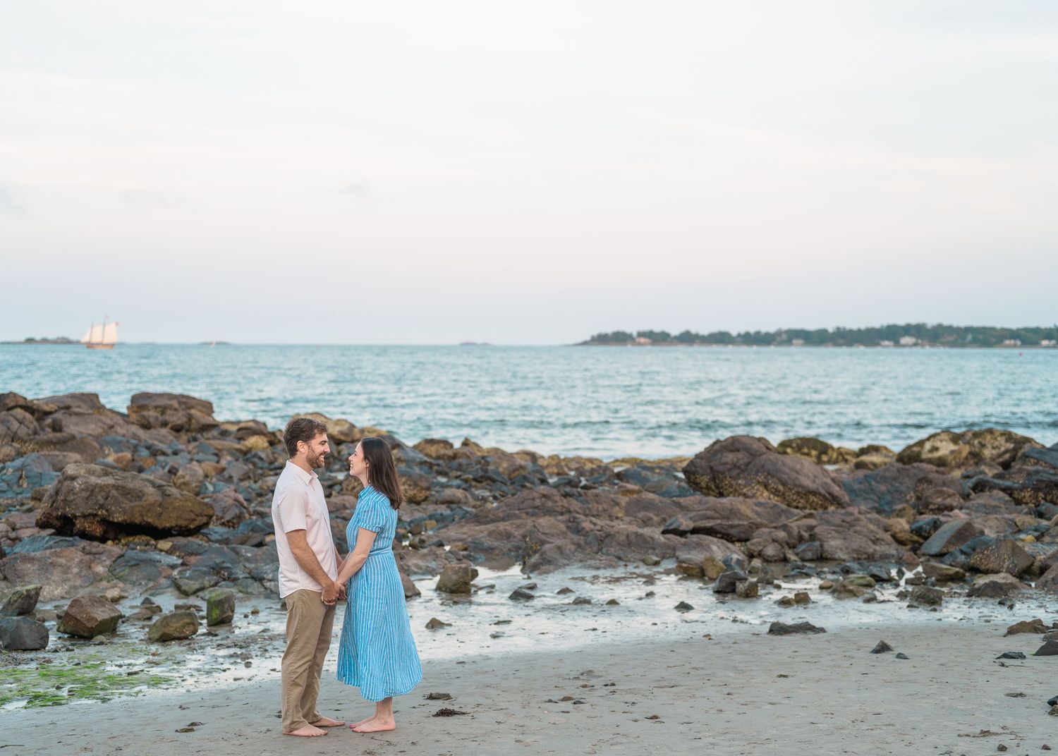 A couple stands on a rocky beach at sunset with sailboats visible in the distance across calm waters.