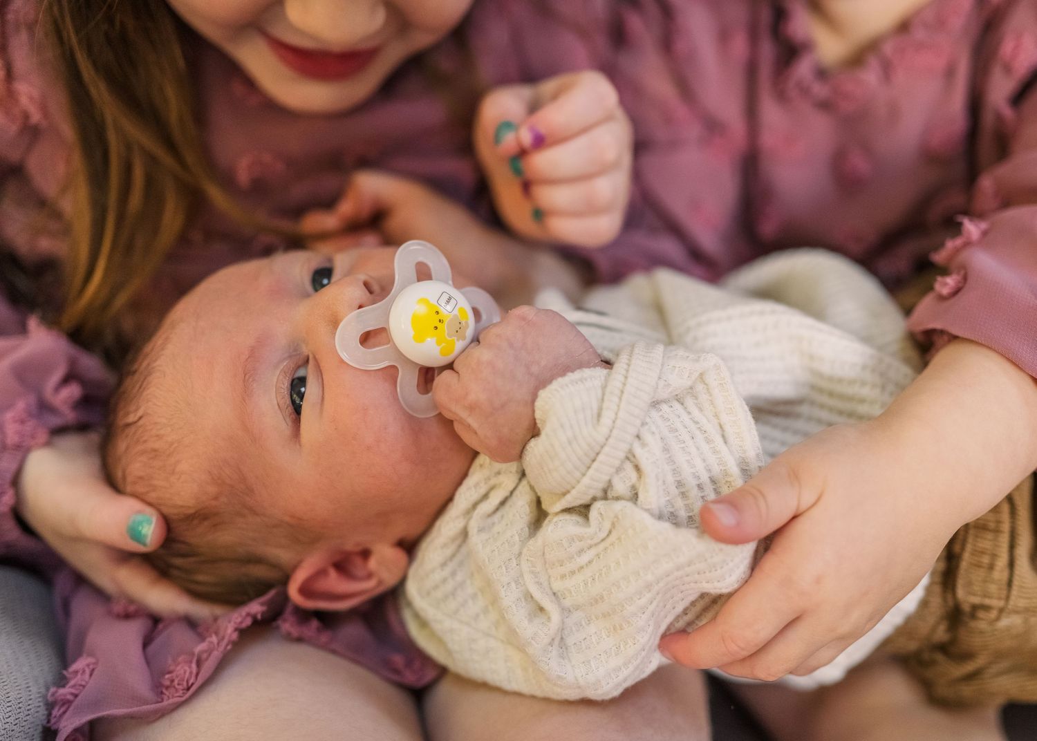 Newborn baby with pacifier surrounded by family members in pink clothing.