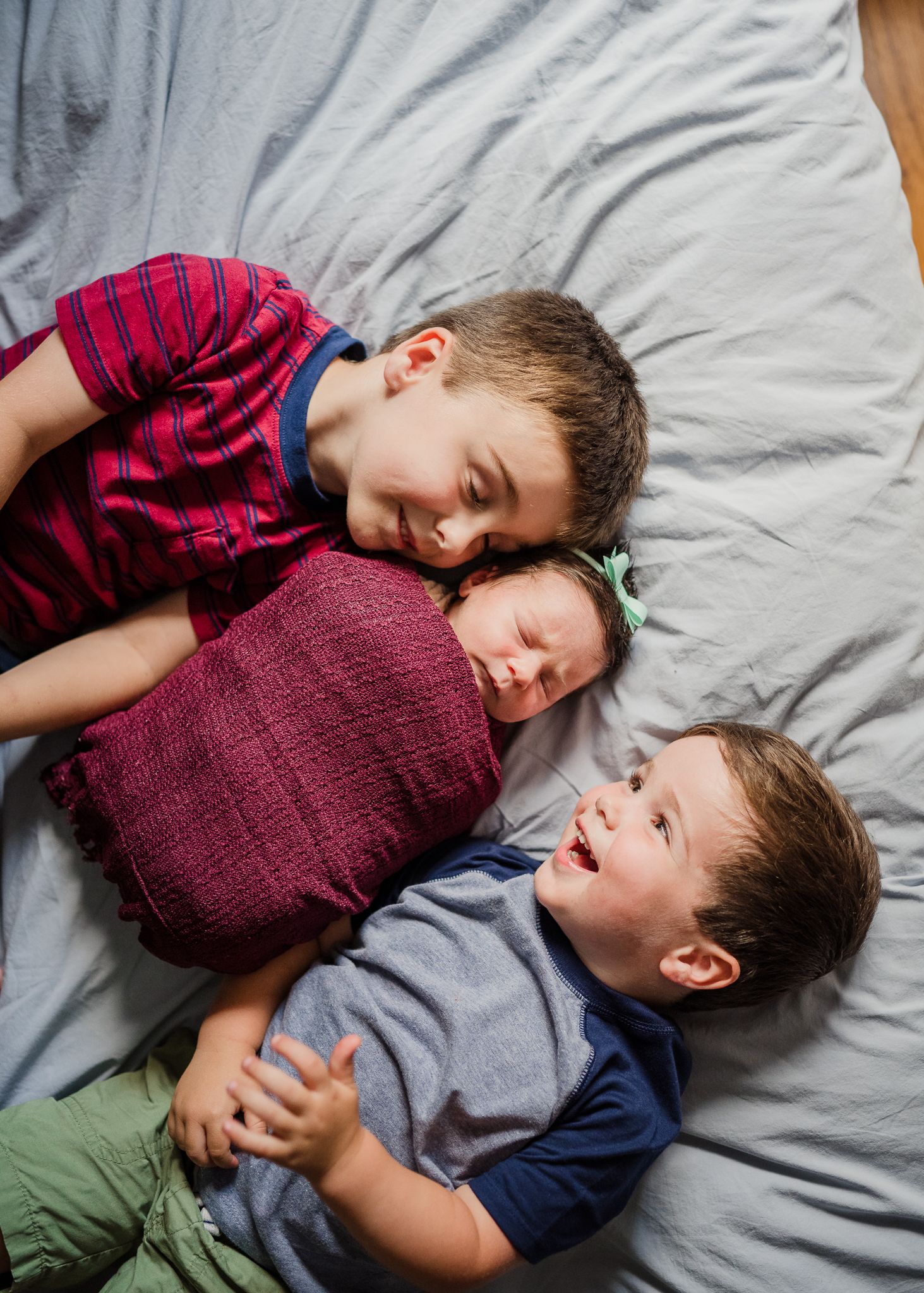 Three children lying together playfully on grey bedding.