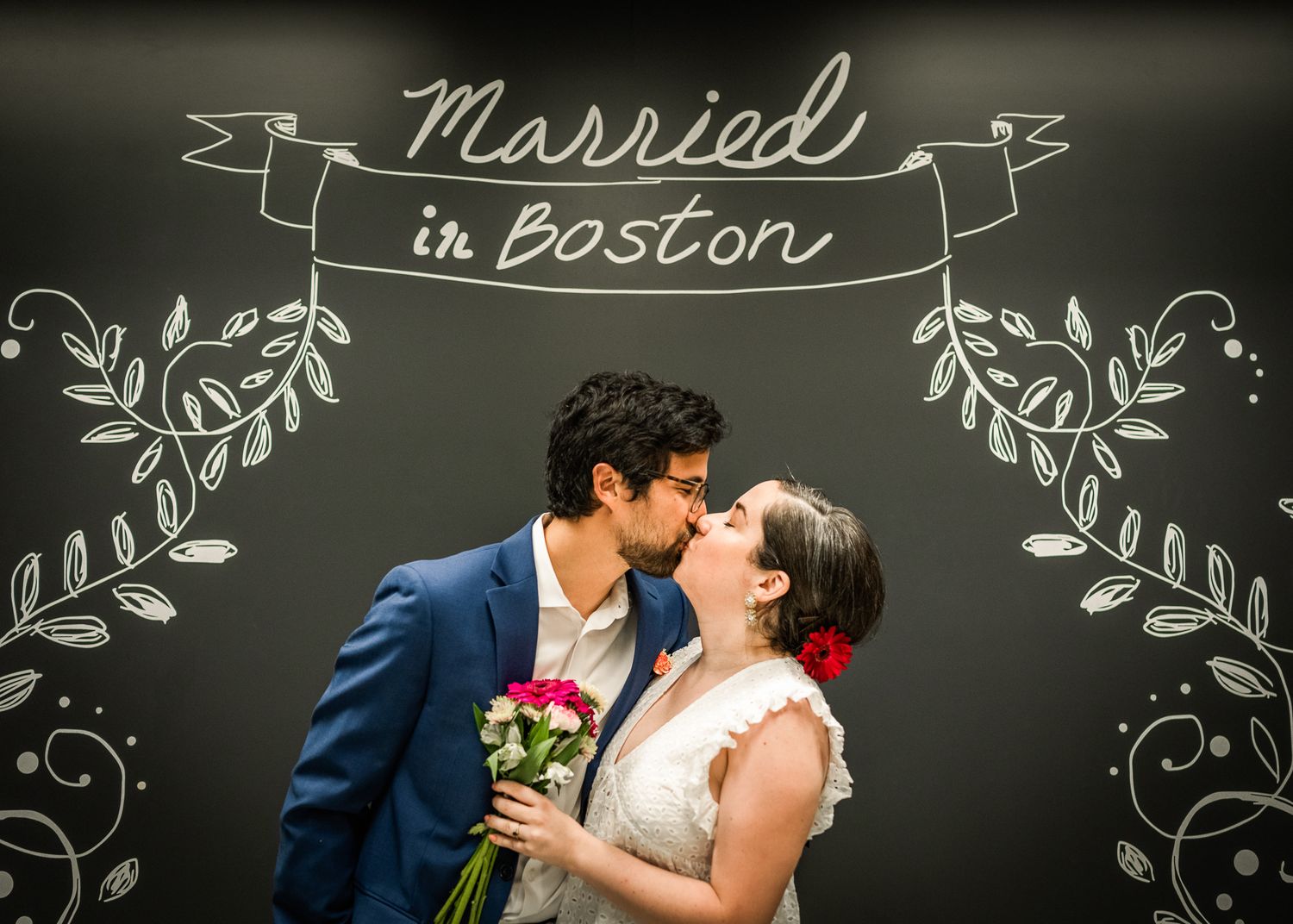 Wedding couple kisses under hand-drawn 'Married in Boston' chalkboard sign with floral details.