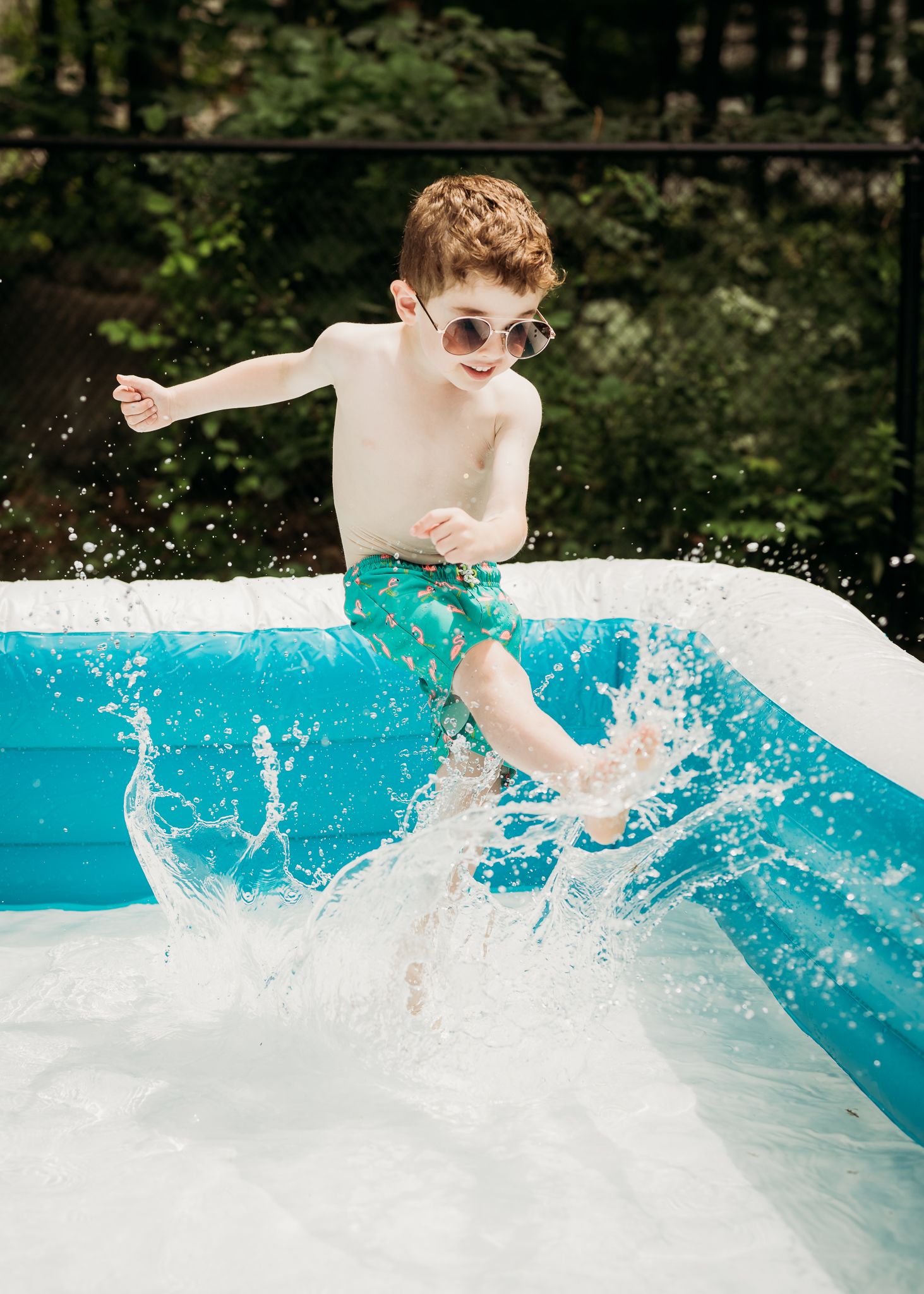 Child in sunglasses and turquoise swimwear splashing into a pool with water spray on a sunny day.