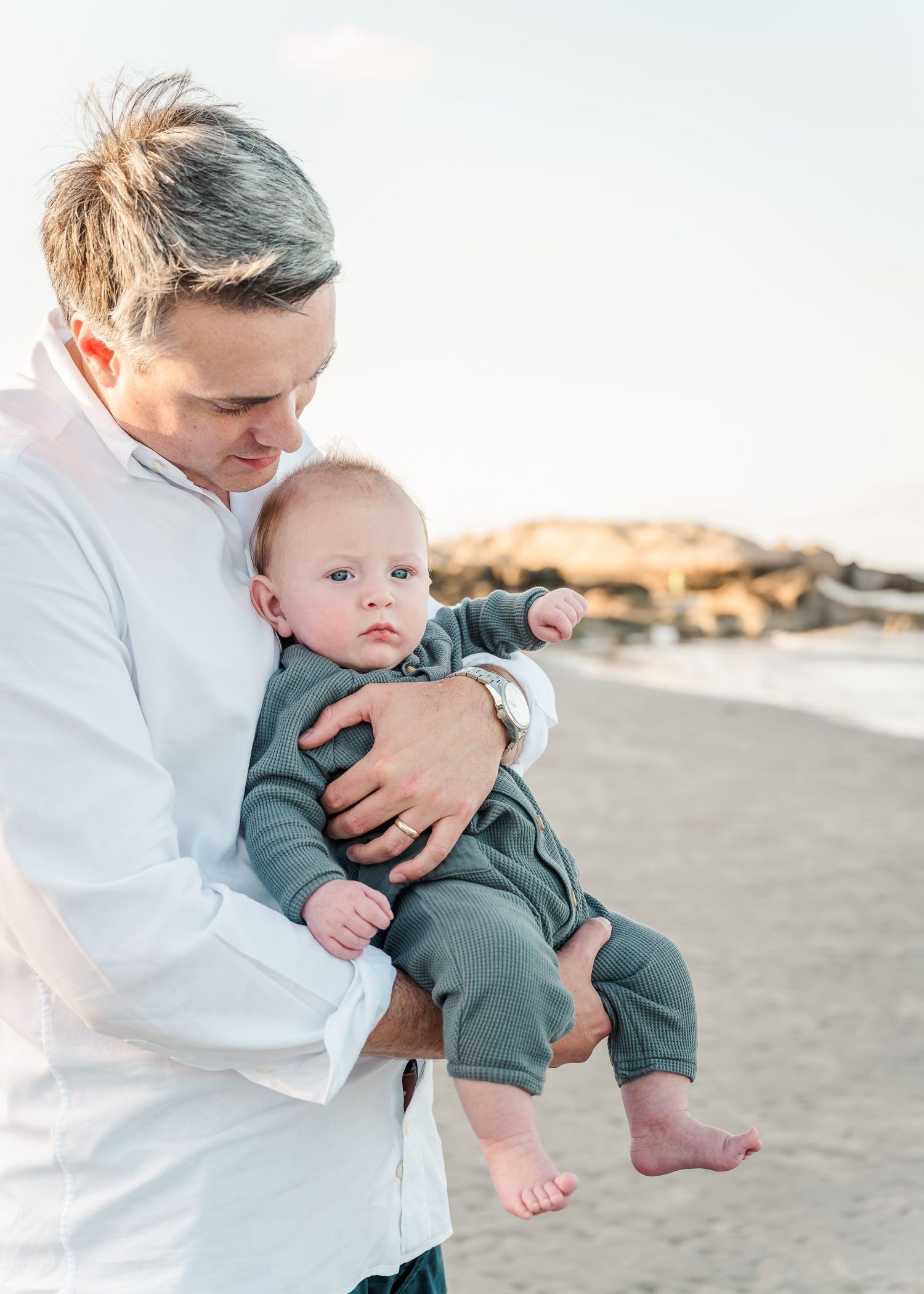 A tender moment captured at the beach with a baby wearing a teal romper being held against the ocean backdrop.