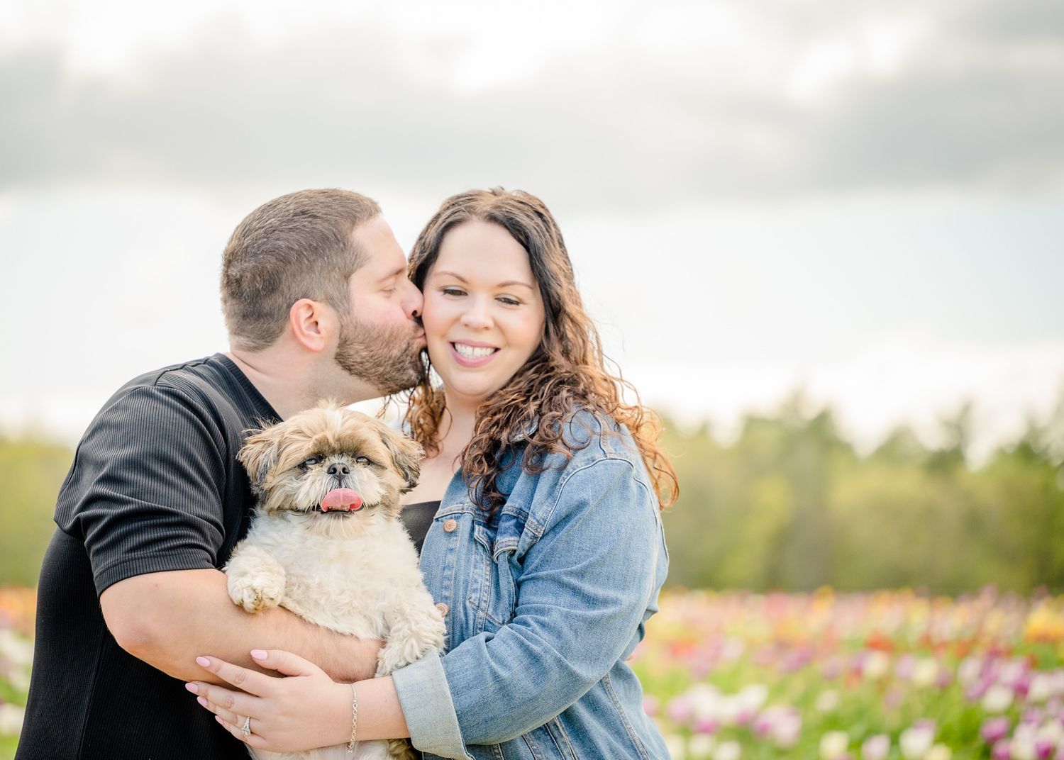Couple holds their small fluffy dog while posing in field of flowers.