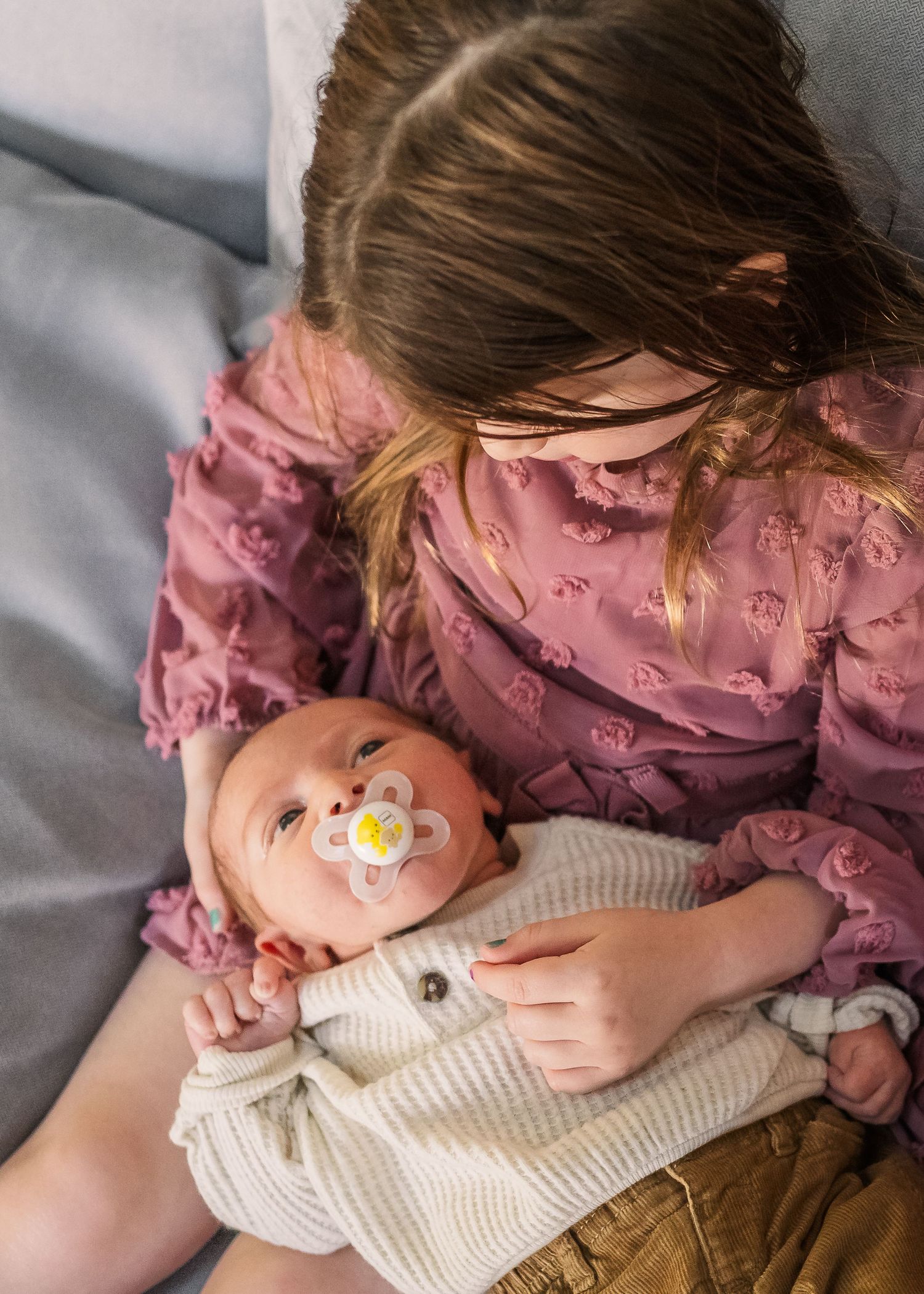 Young child in pink dress cuddling with baby wearing pacifier on grey couch.