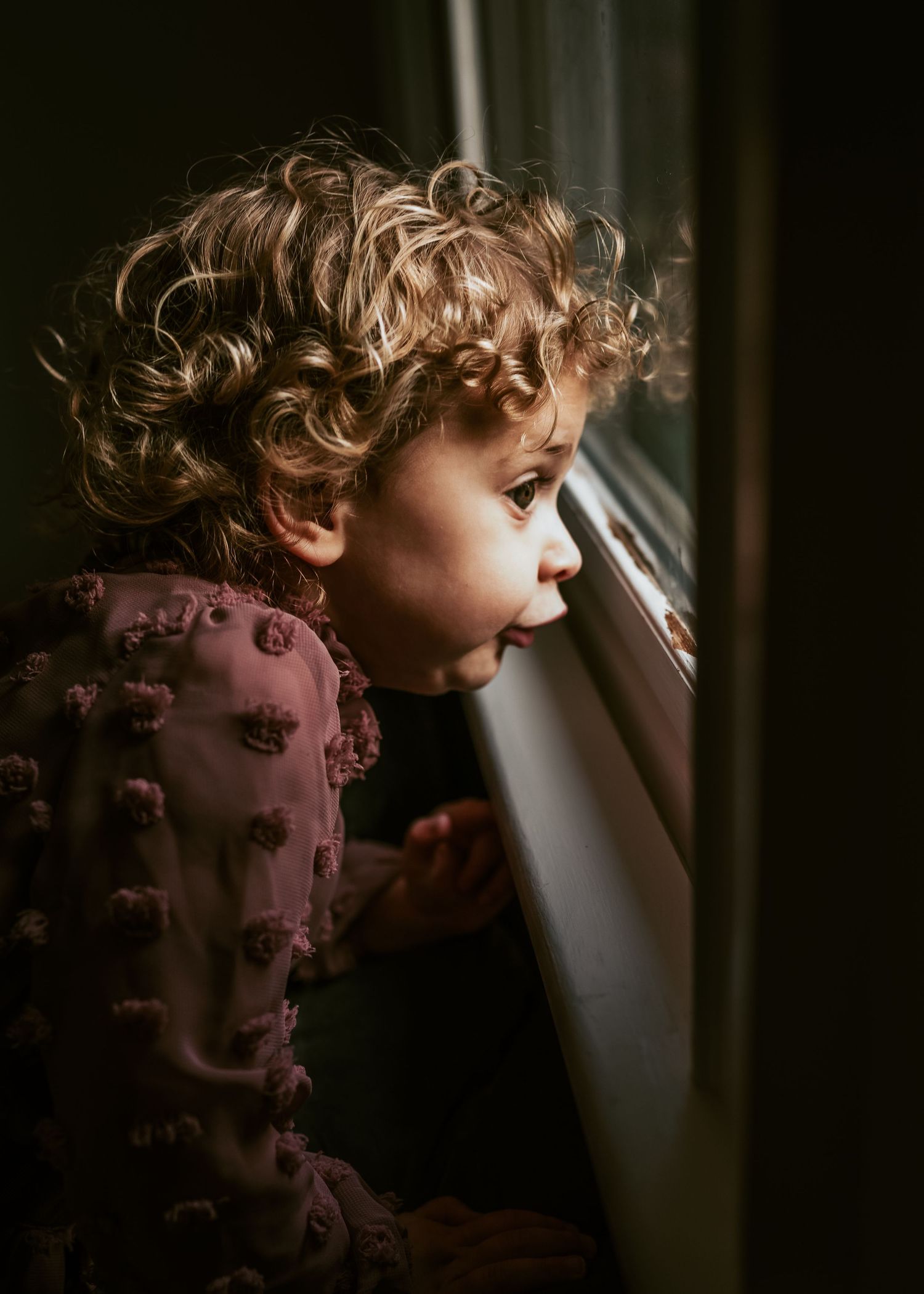 A toddler in pink dress looking curiously through a window in moody lighting.