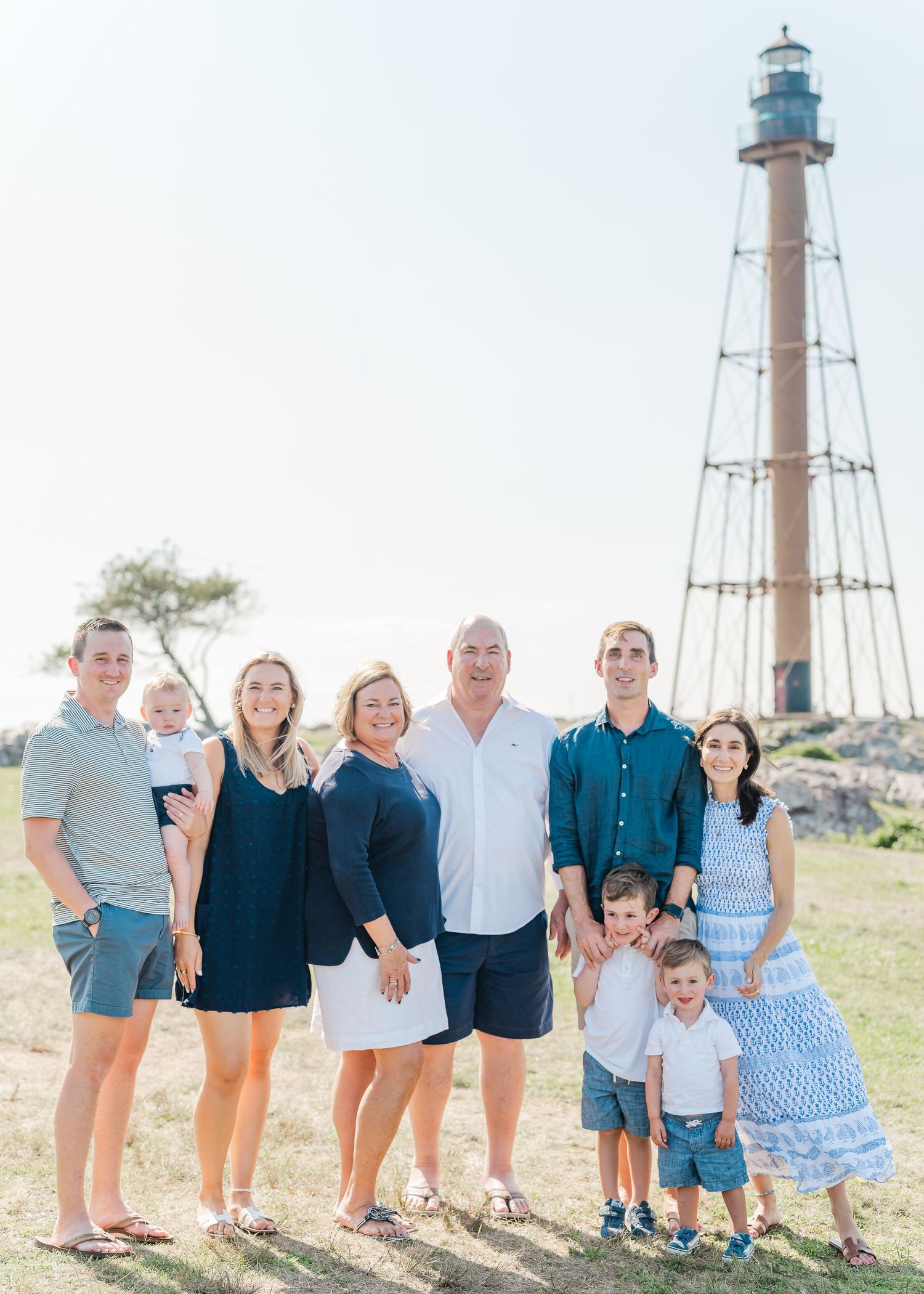 A family poses together on a grassy hill with a tall lighthouse structure visible in the background on a sunny day.