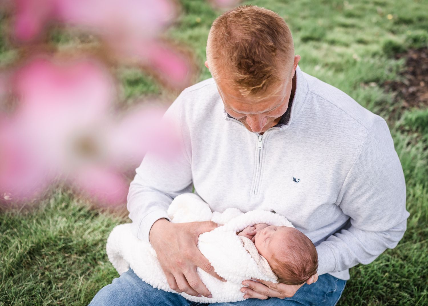 Parent cradles sleeping newborn outdoors with pink blossoms in background.