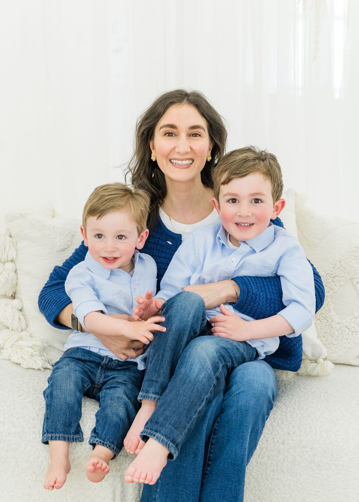Portrait of a loving family wearing coordinating blue and denim outfits sitting together barefoot on white couch.