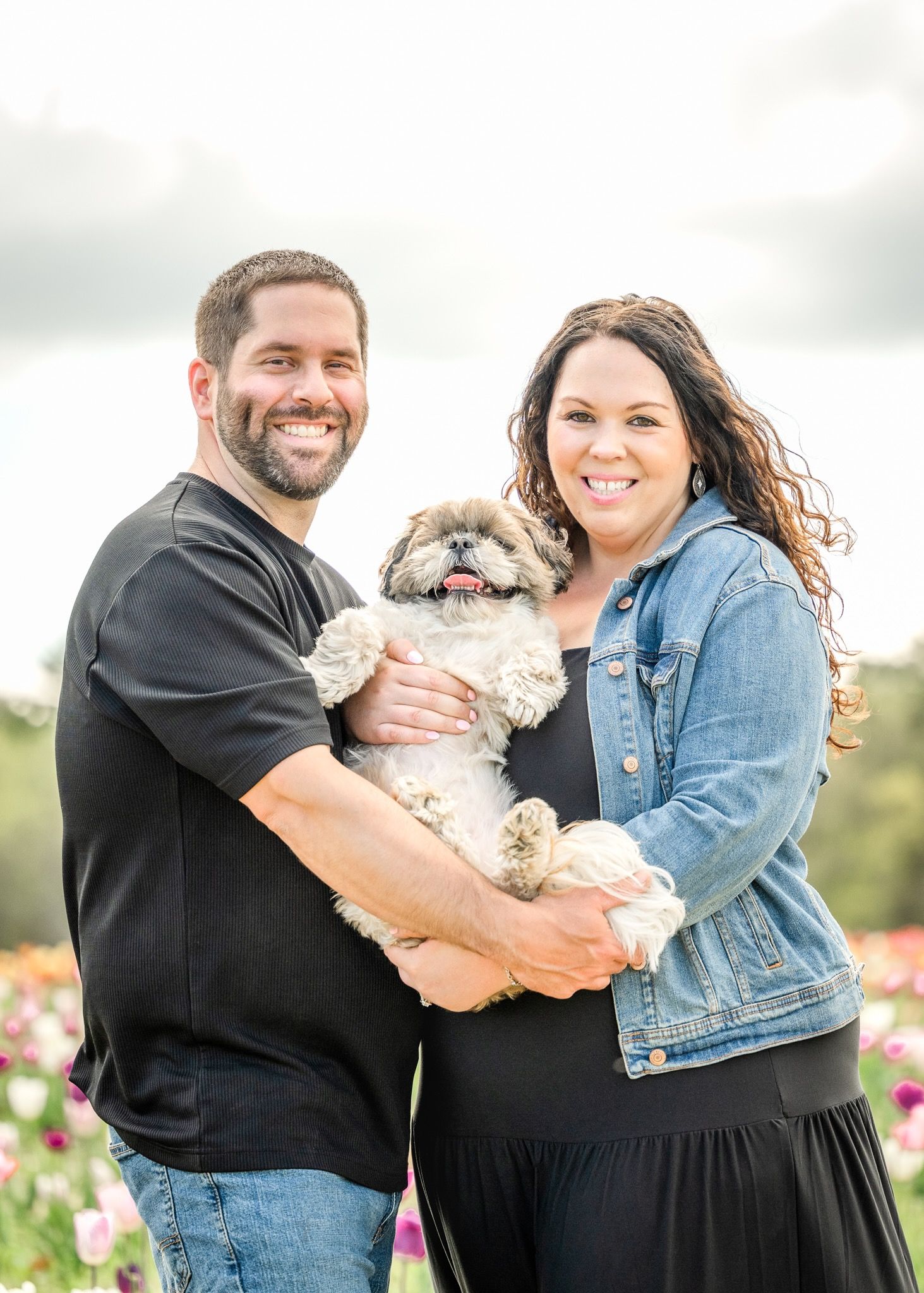 Couple in casual attire holding their happy Shih Tzu dog in flowering field.