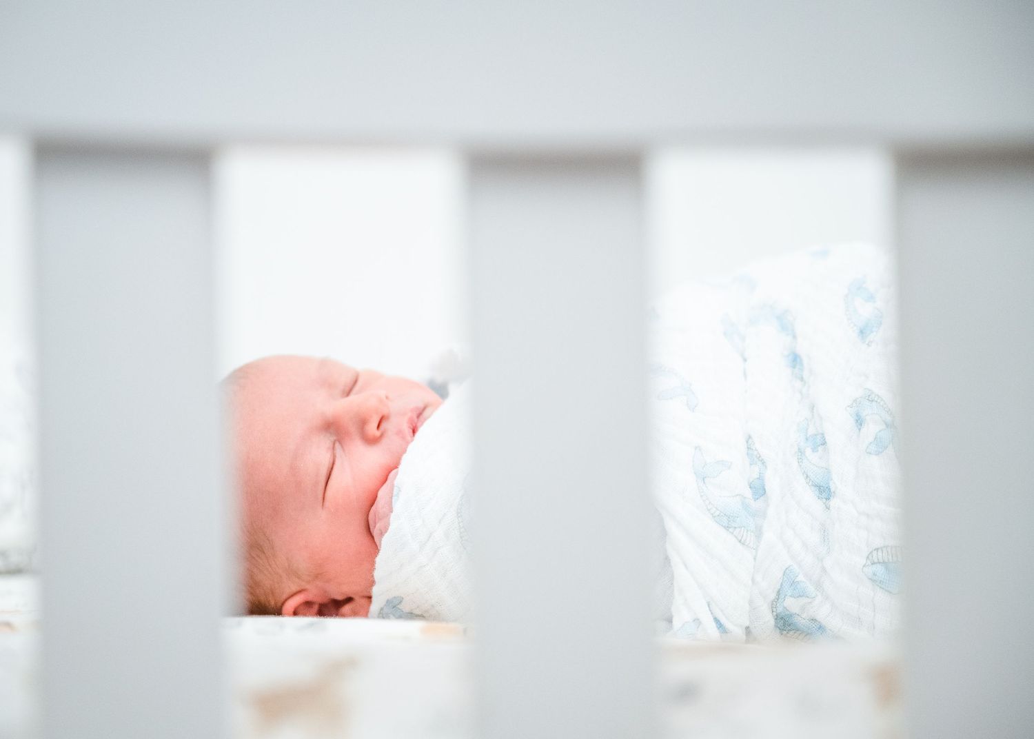 Sleeping newborn viewed through white crib slats in bright nursery setting.