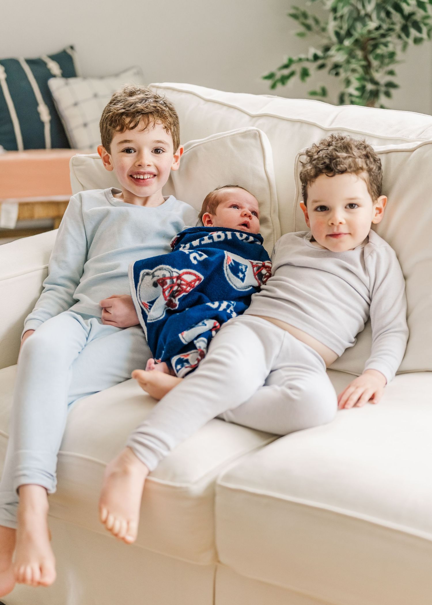 Three brothers lounging on a white couch, with a newborn baby in blue pajamas nestled between his siblings.