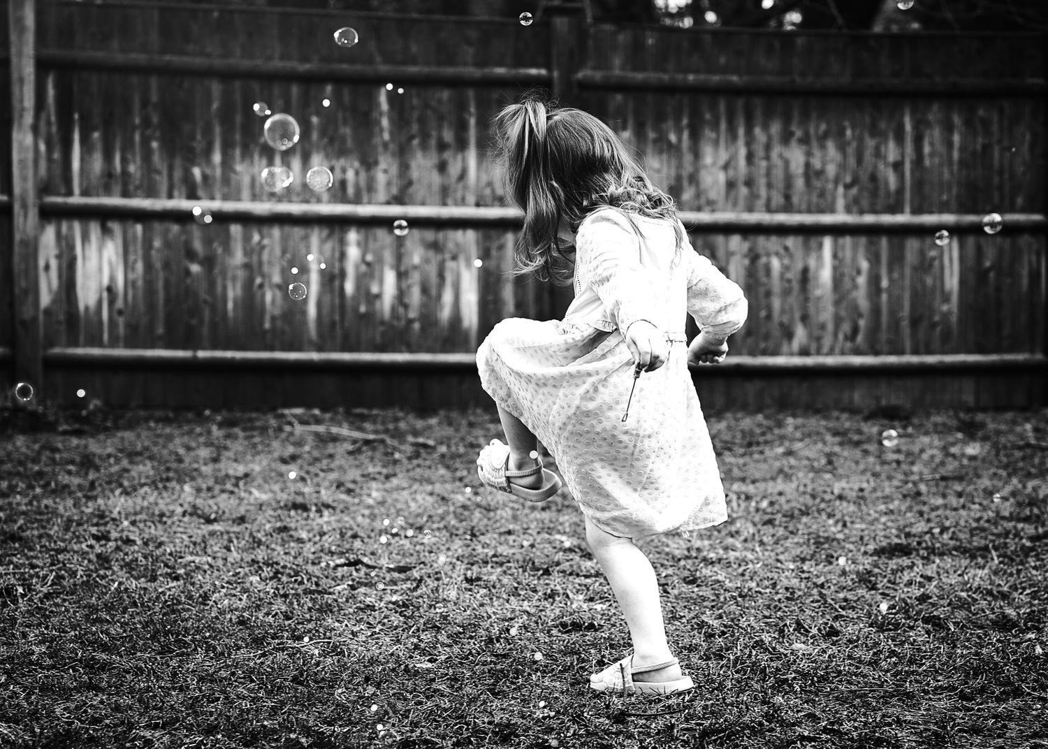 A child in a white dress running and playing in a backyard, captured in black and white.