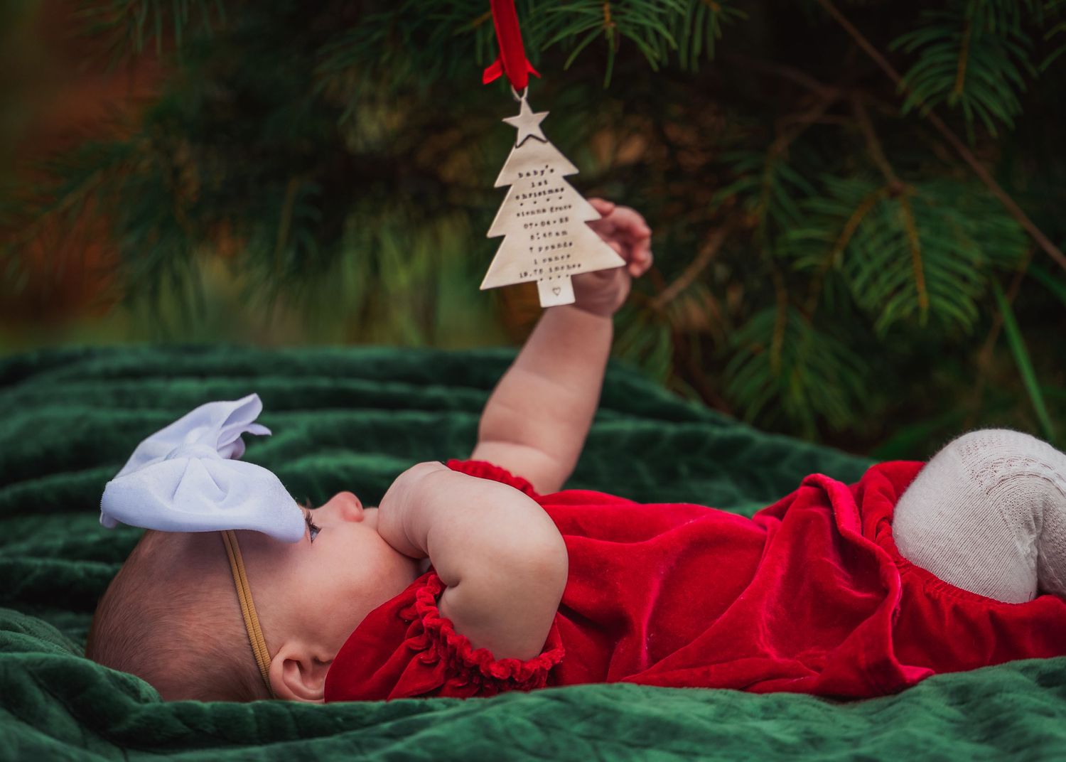 Baby in red outfit lying on green blanket reaching for Christmas tree ornament.