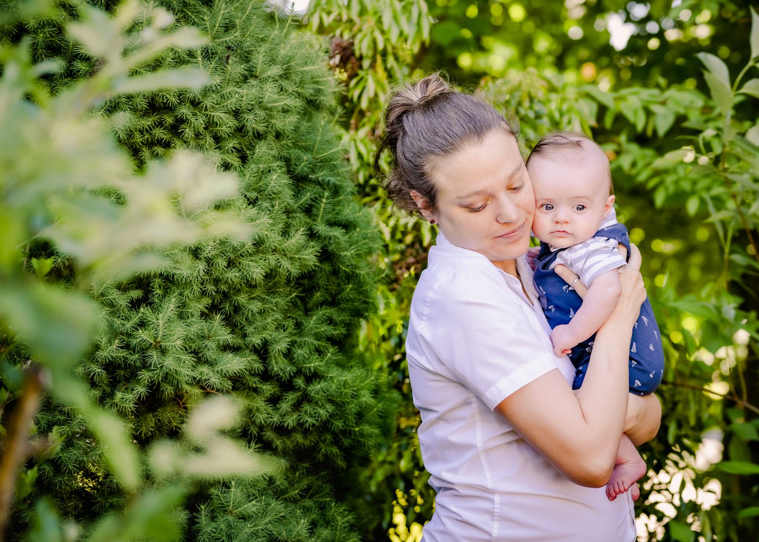 A parent holds a small baby wearing blue against a backdrop of green garden foliage.