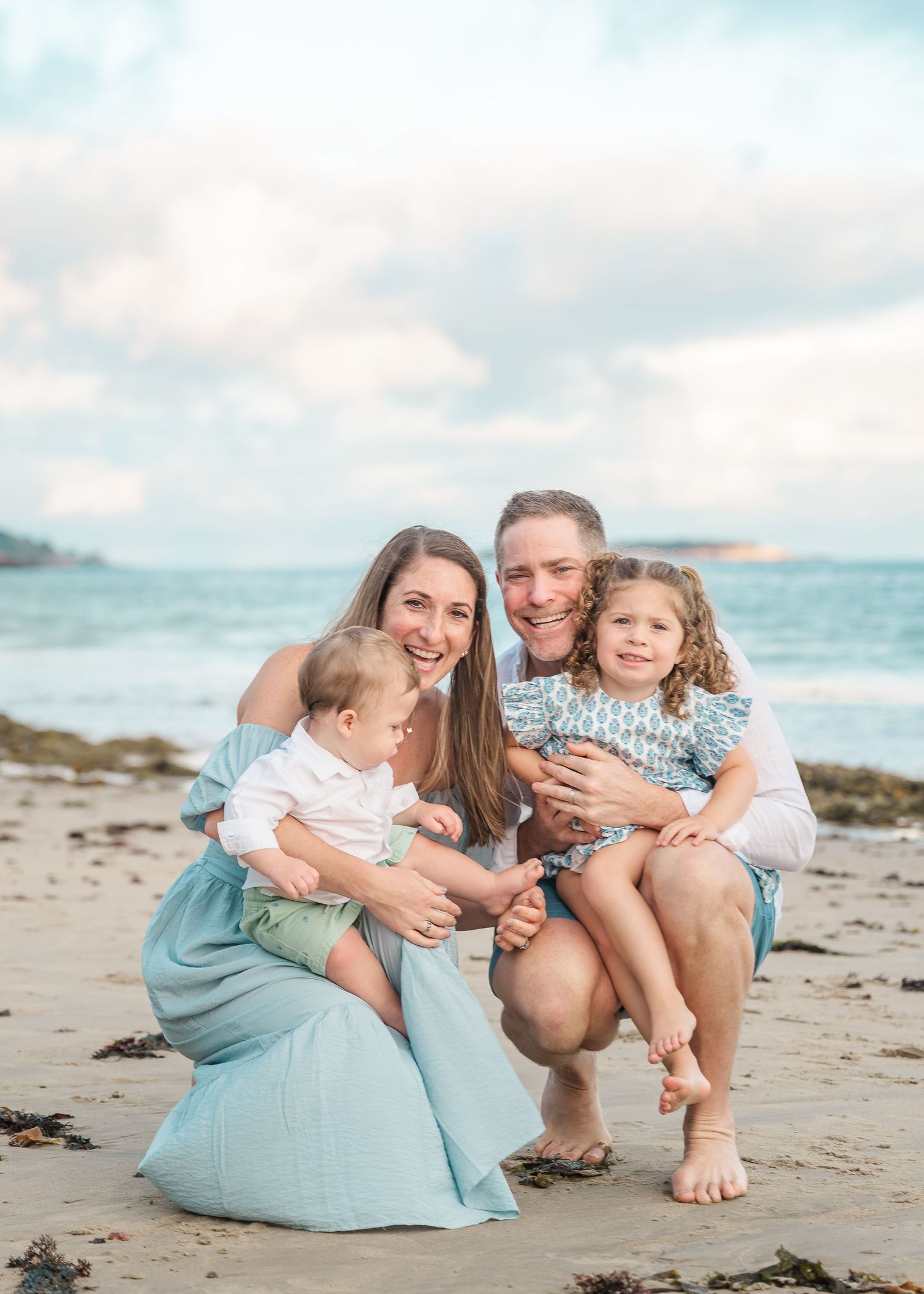 A happy family in coordinating blue outfits poses together on a scenic beach with ocean waves behind them.