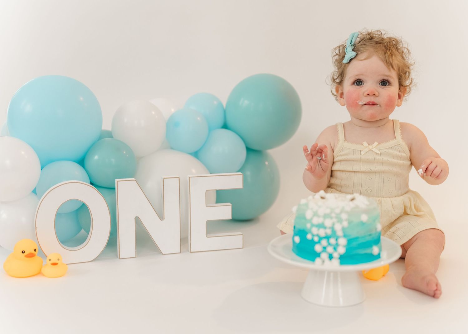 Cake smash. Baby in white onesie sits with blue balloons and white ONE letters during first birthday cake smash photo session.