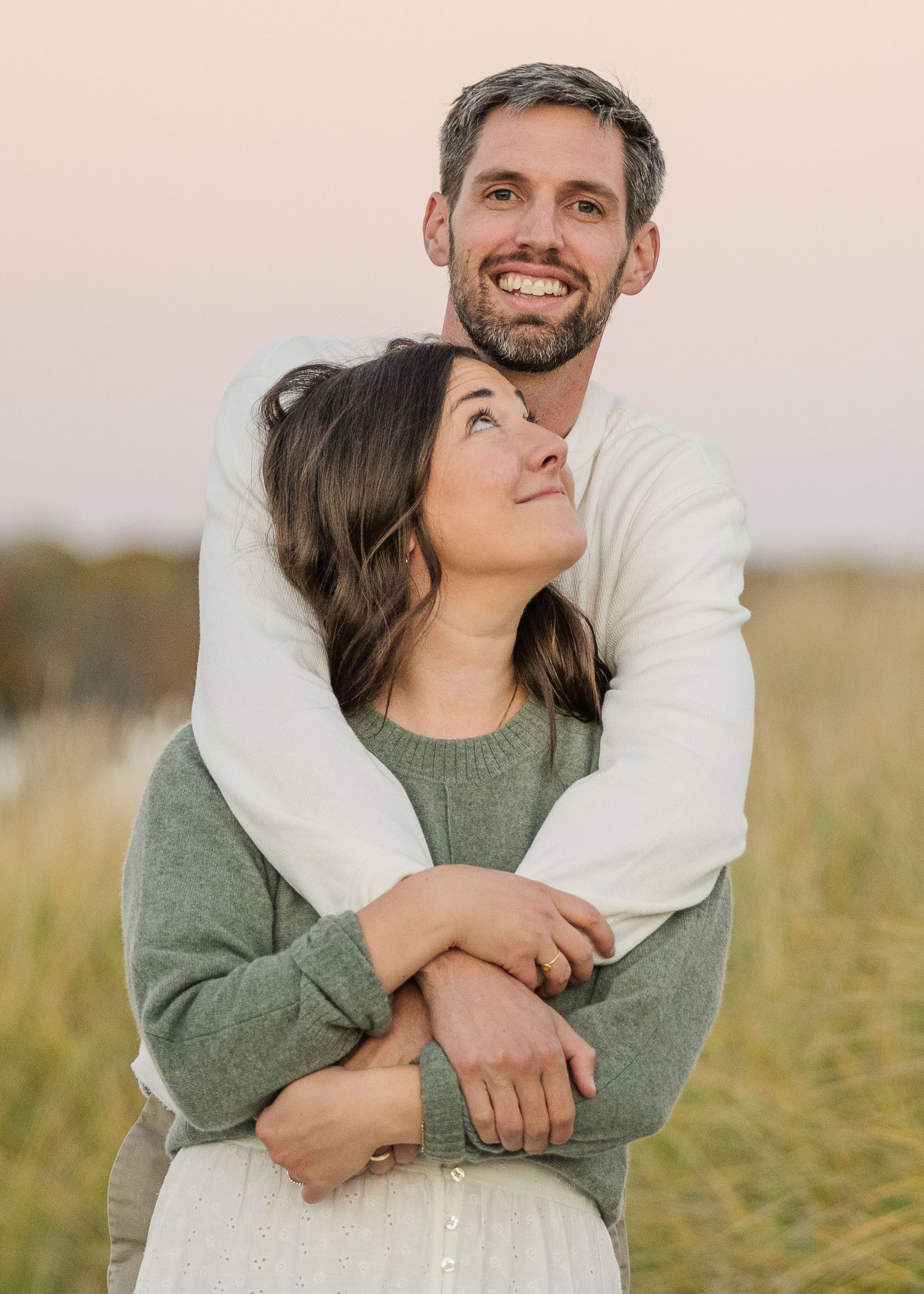 A couple embraces lovingly in a sunlit field wearing green and white casual attire.