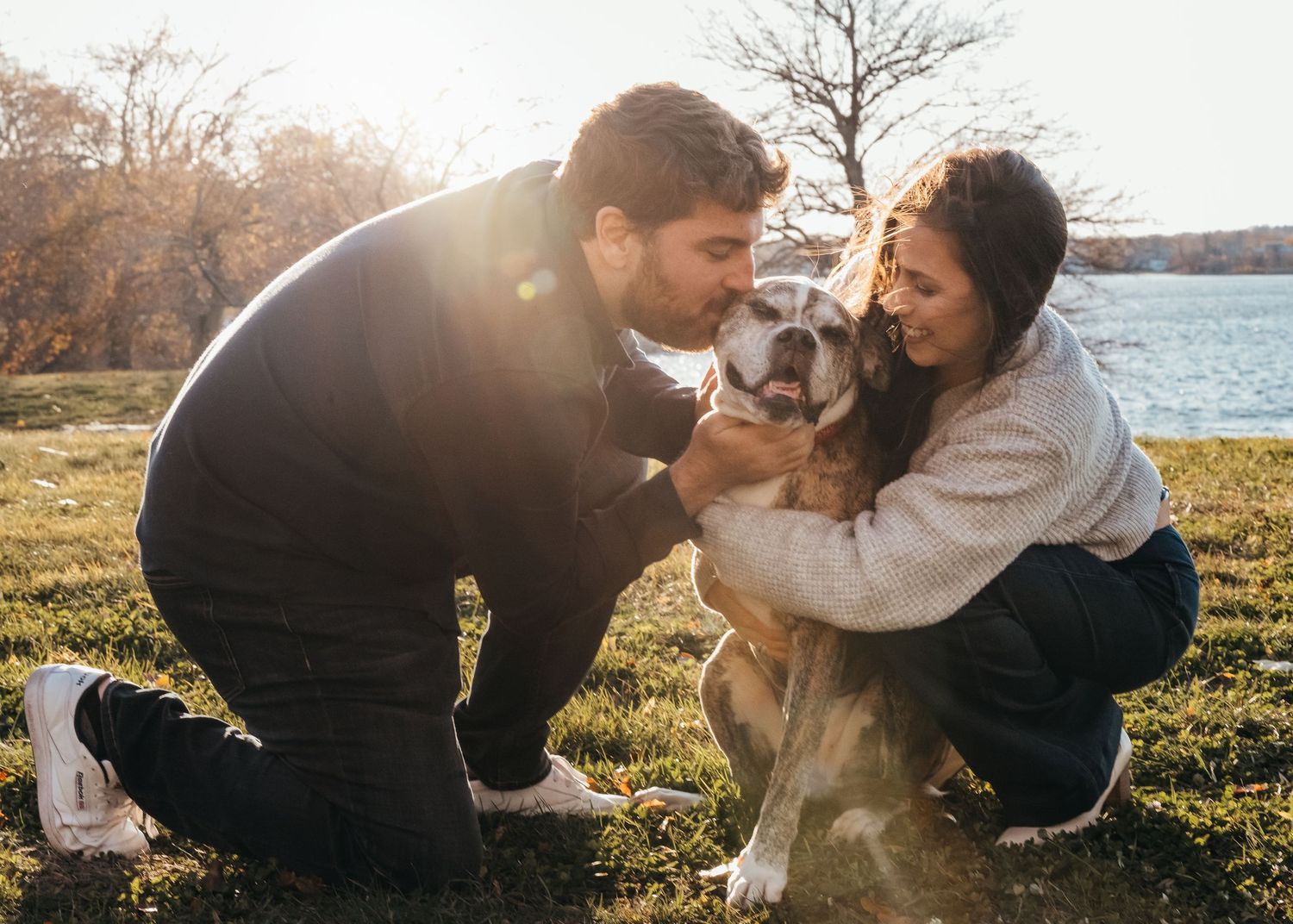 A couple shares a joyful moment with their husky dog in a scenic park by the water on a sunny autumn day.