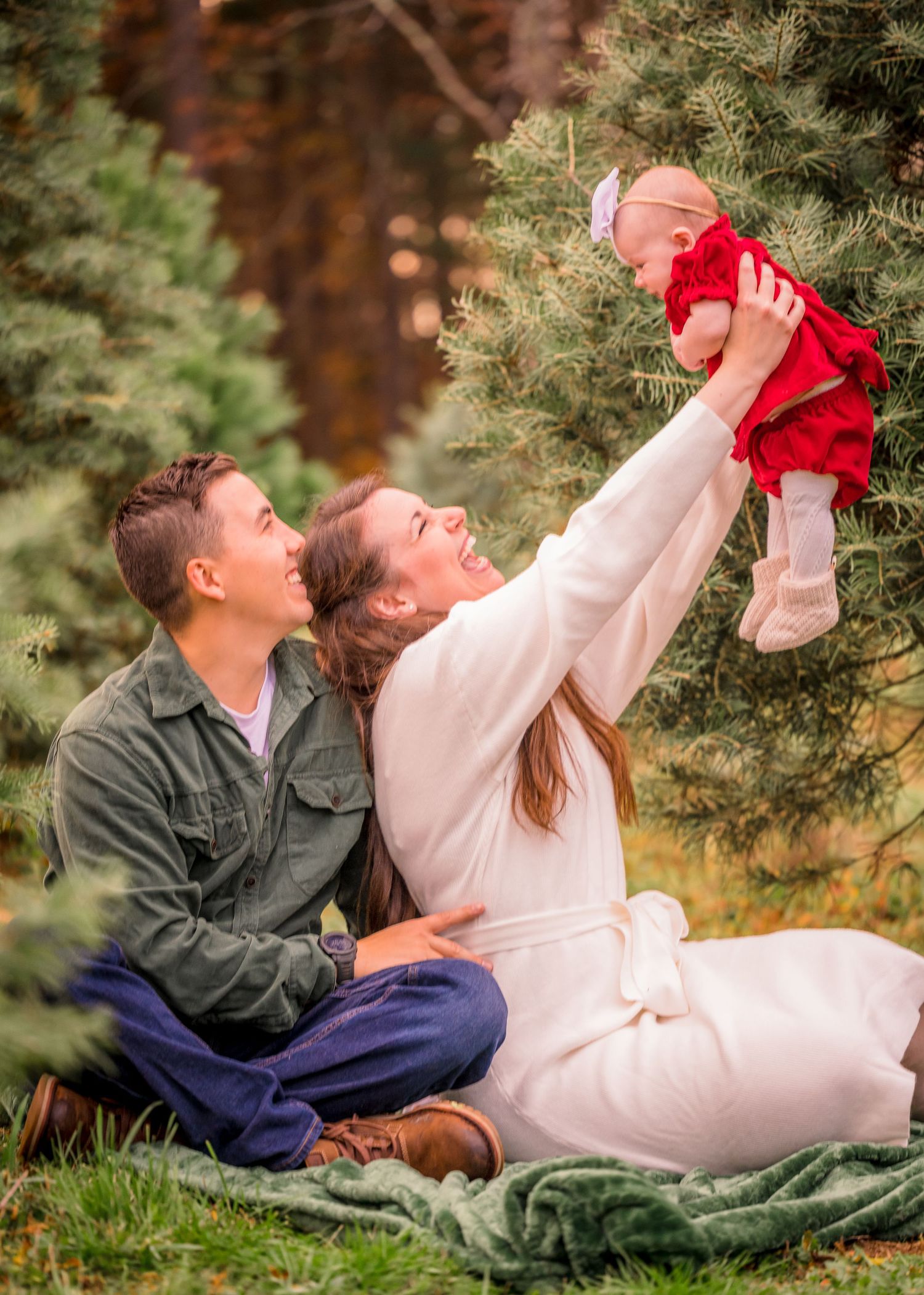 Joyful family moment captured outdoors with baby being lifted up among pine trees.