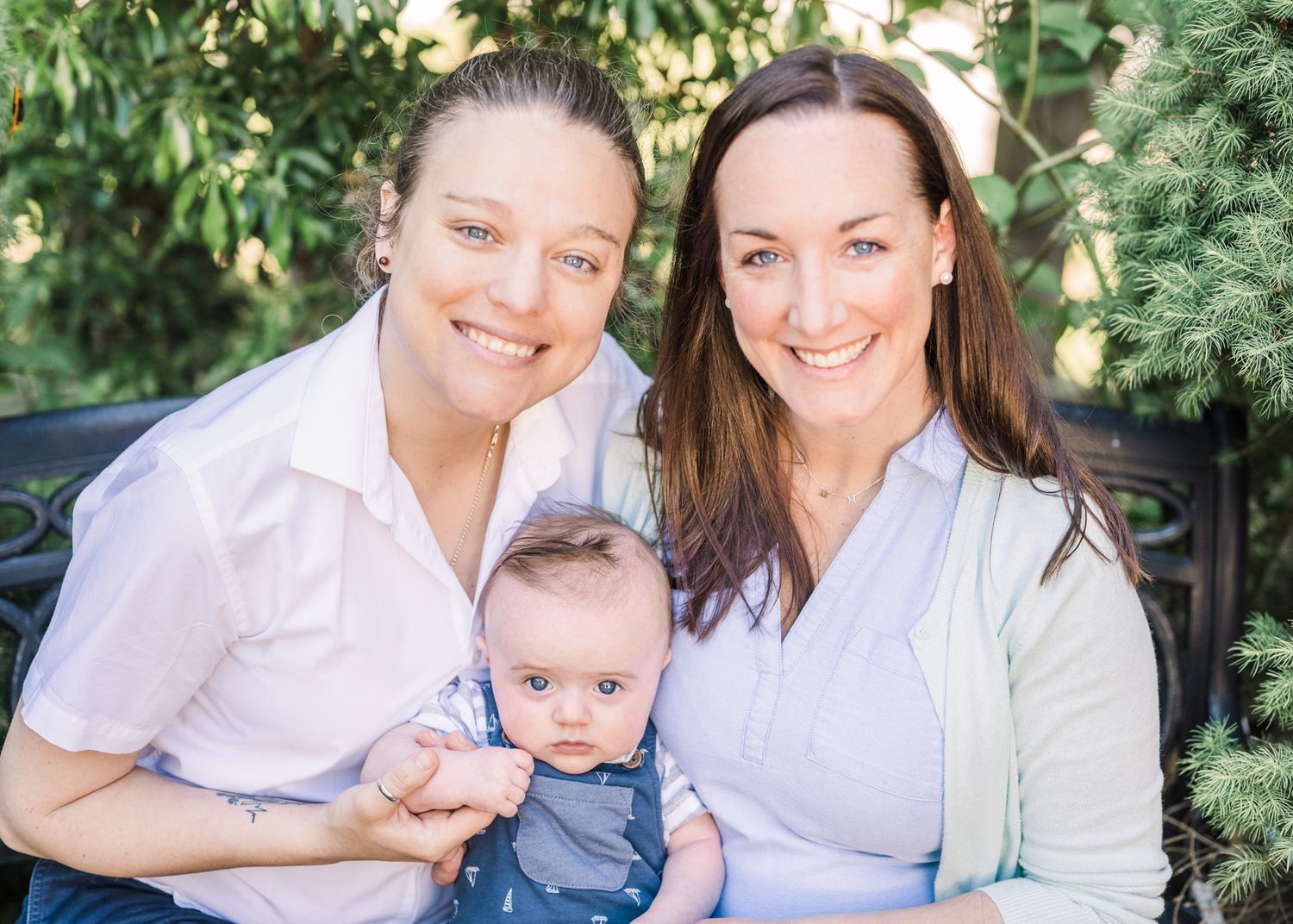 Family portrait outdoors featuring a baby in blue overalls with evergreen trees in the background.