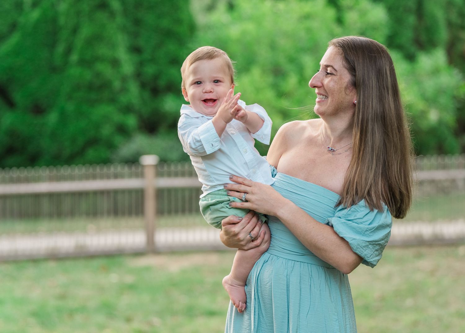 A mother in a light blue dress holds a smiling baby outdoors in a green garden setting.
