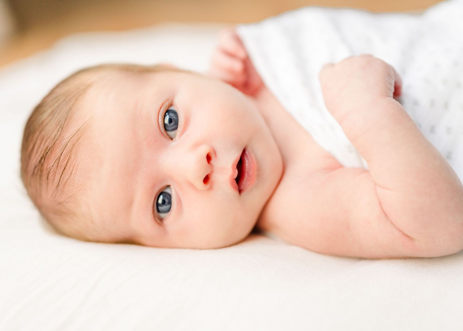 Close up of awake baby with bright blue eyes lying on white bedding.