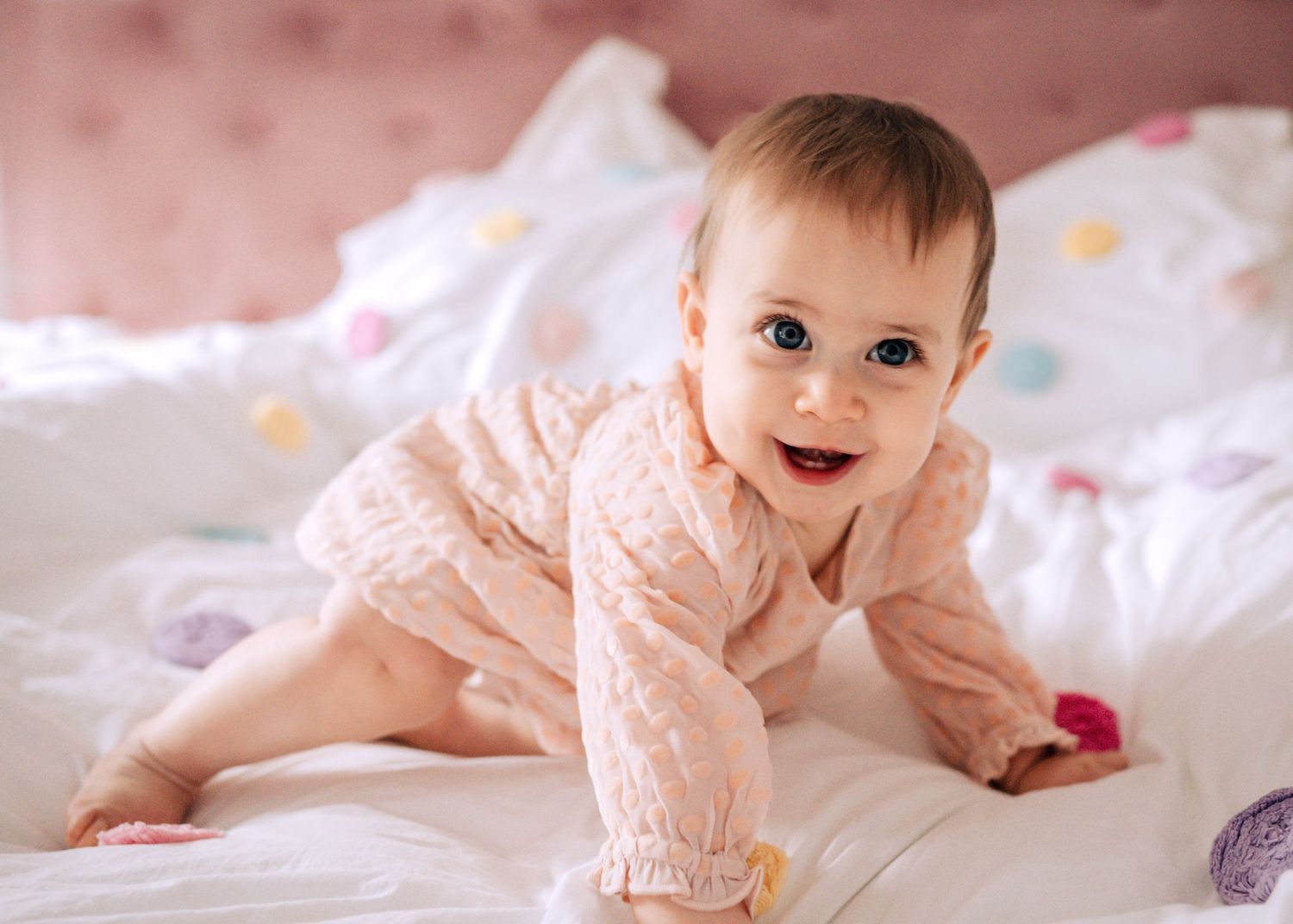 A smiling baby in a pink polka dot onesie crawls on white bedding.