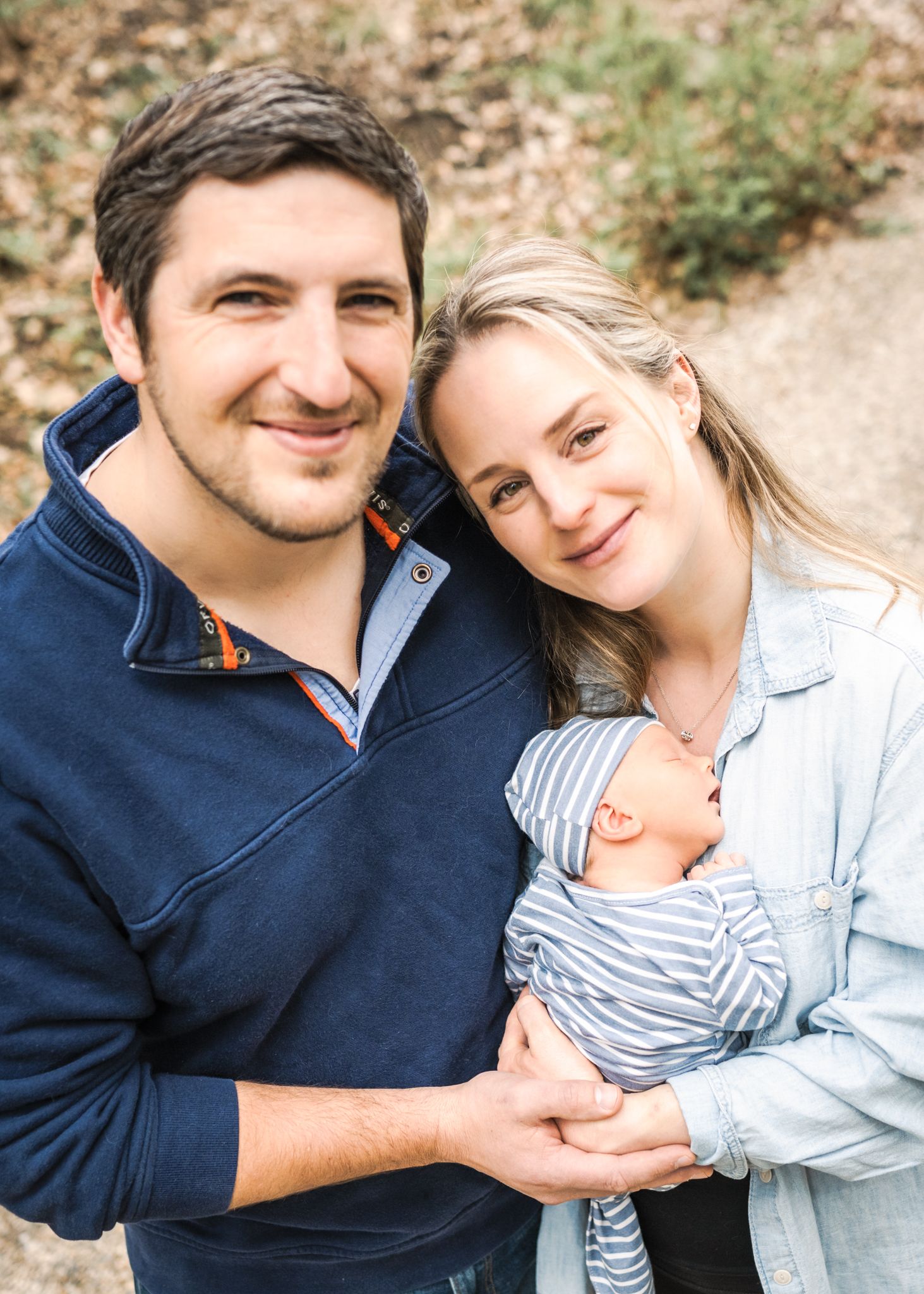 Outdoor family portrait with infant in striped outfit on sunny day.
