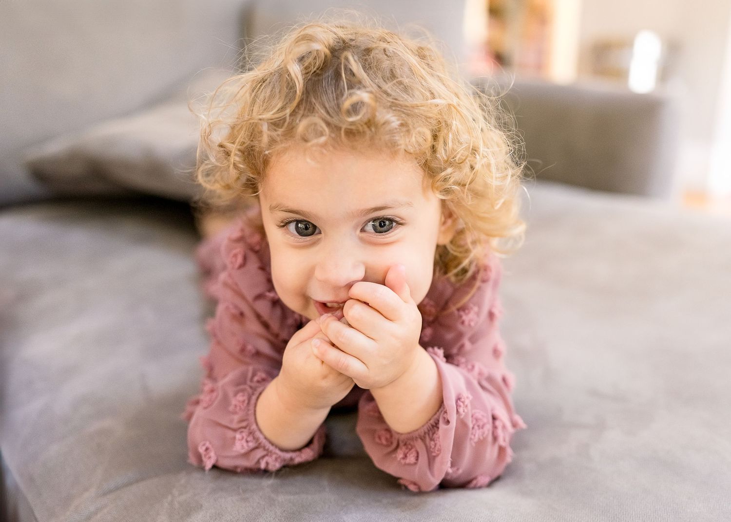 Young child in pink sweater lying on grey couch with hands covering mouth.