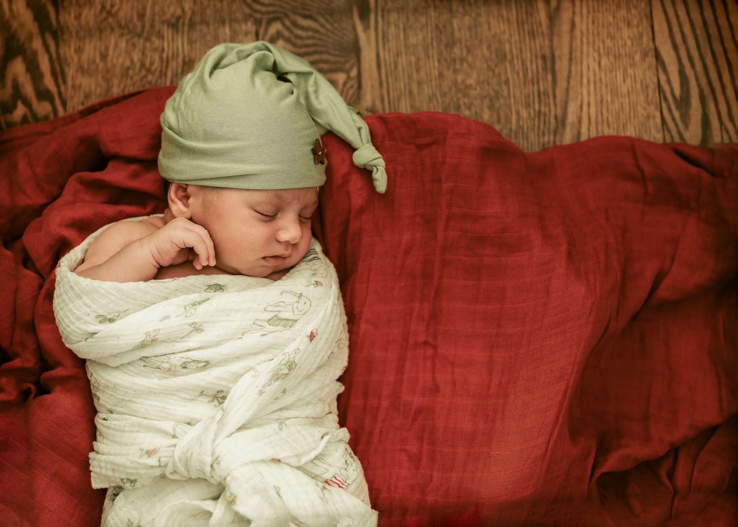 Baby sleeping peacefully in white swaddle and green hat on red cushions.