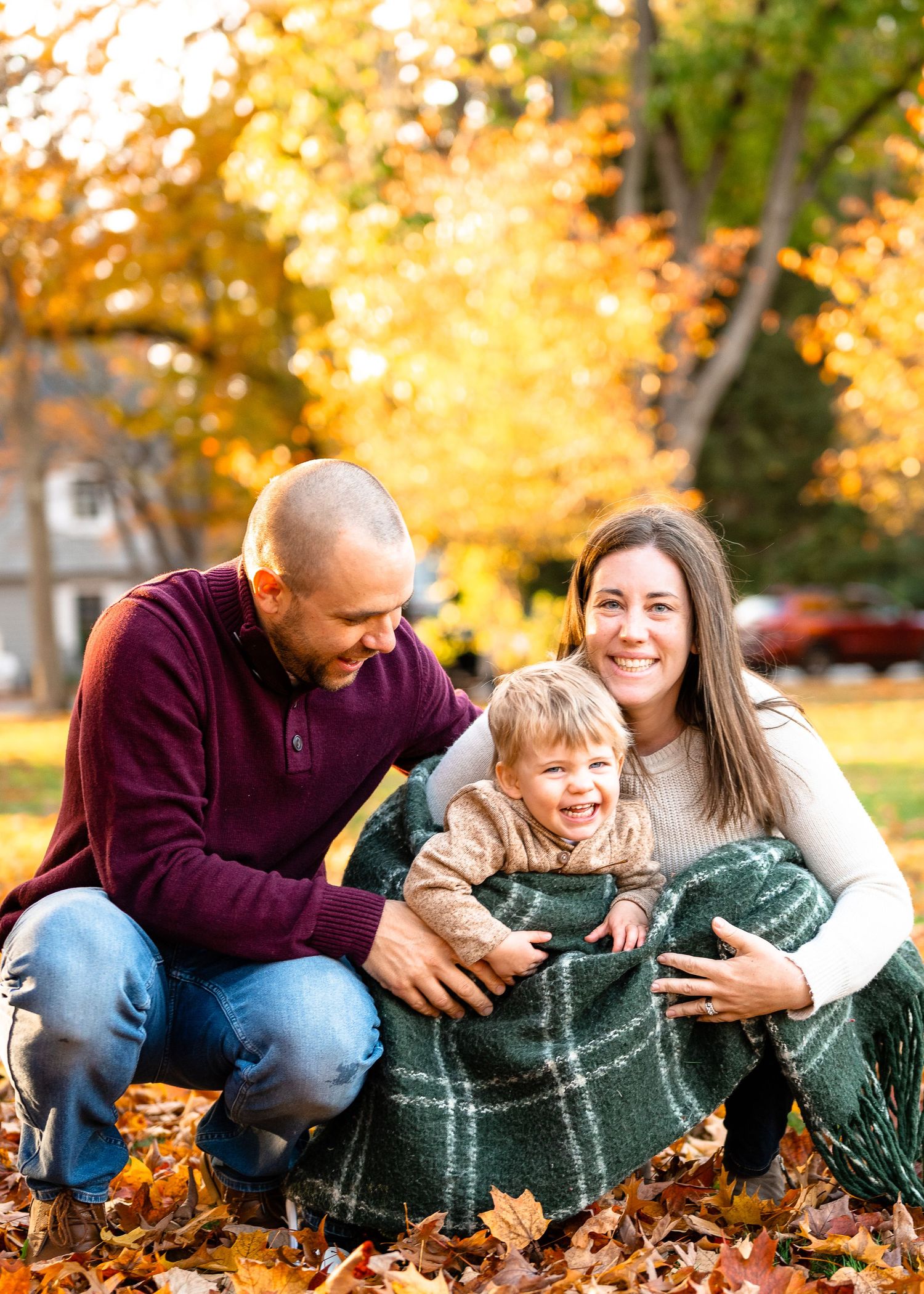 A family shares a tender moment while wrapped in a cozy blanket among autumn leaves.