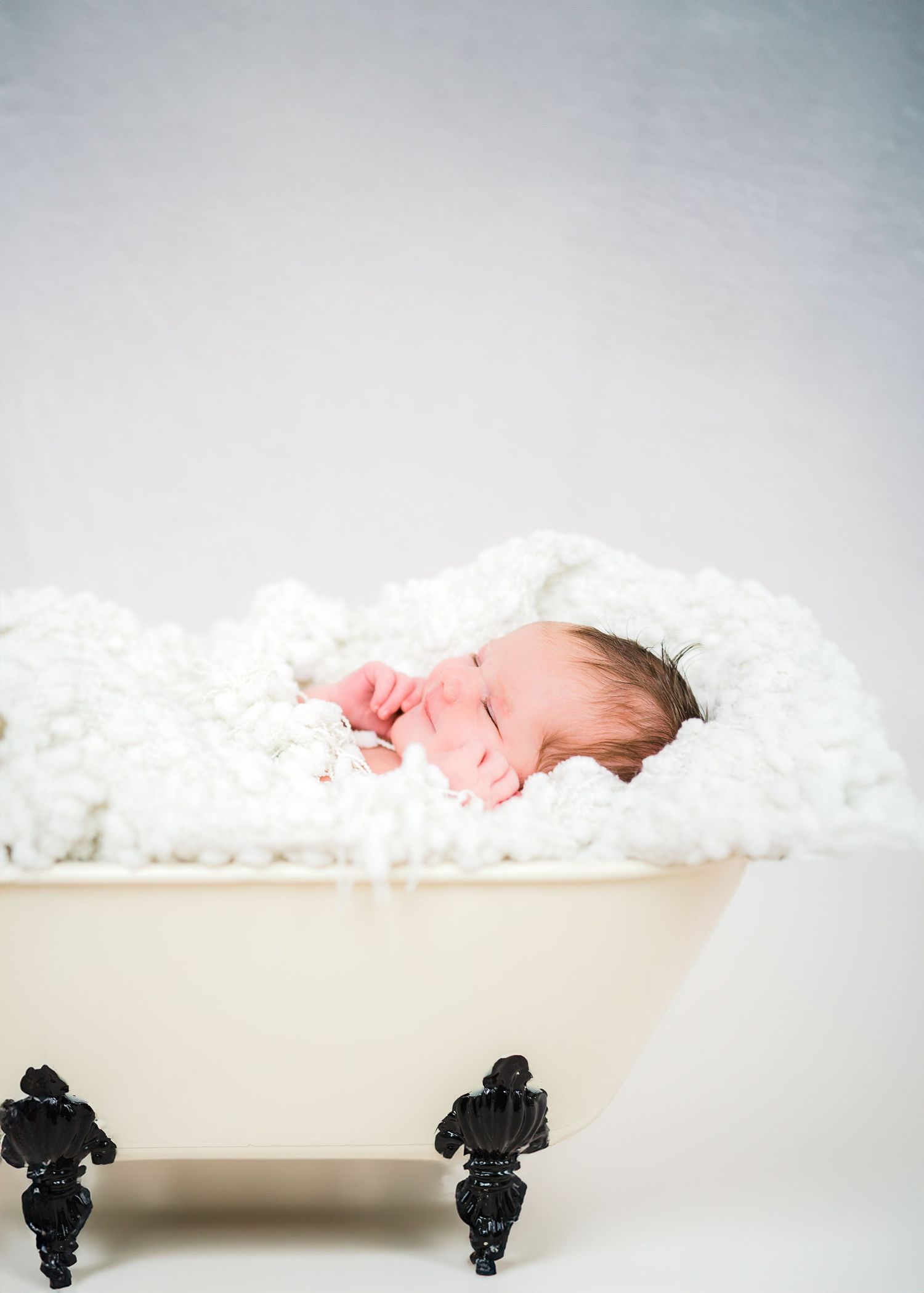 A sleeping newborn rests peacefully in a vintage white clawfoot bathtub filled with soft white blankets.