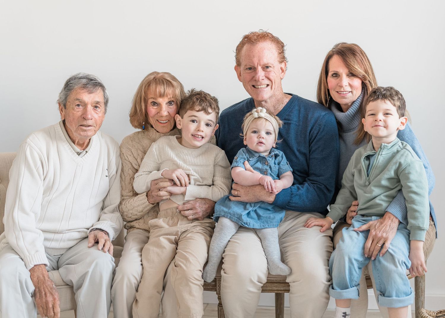 A multi-generational family wearing coordinated neutral-toned outfits sits together on a couch for a portrait session.