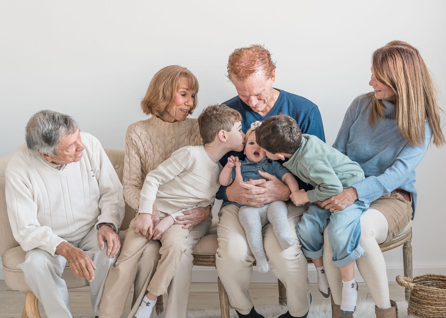 Multi-generational family in neutral colors sitting together on white bench showing affection and joy.