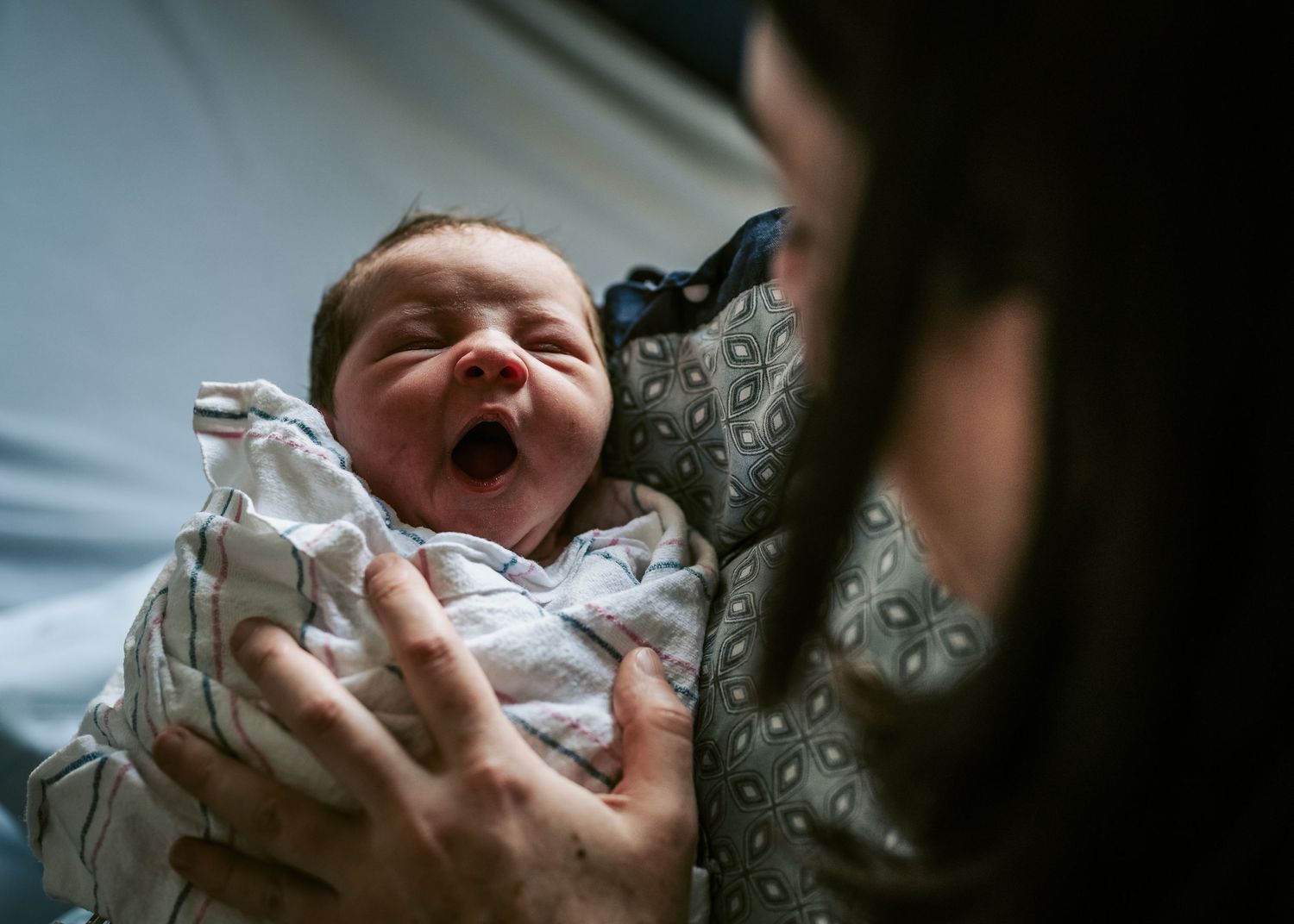 An adorable newborn baby yawns while being cradled in a white blanket during a tender moment.