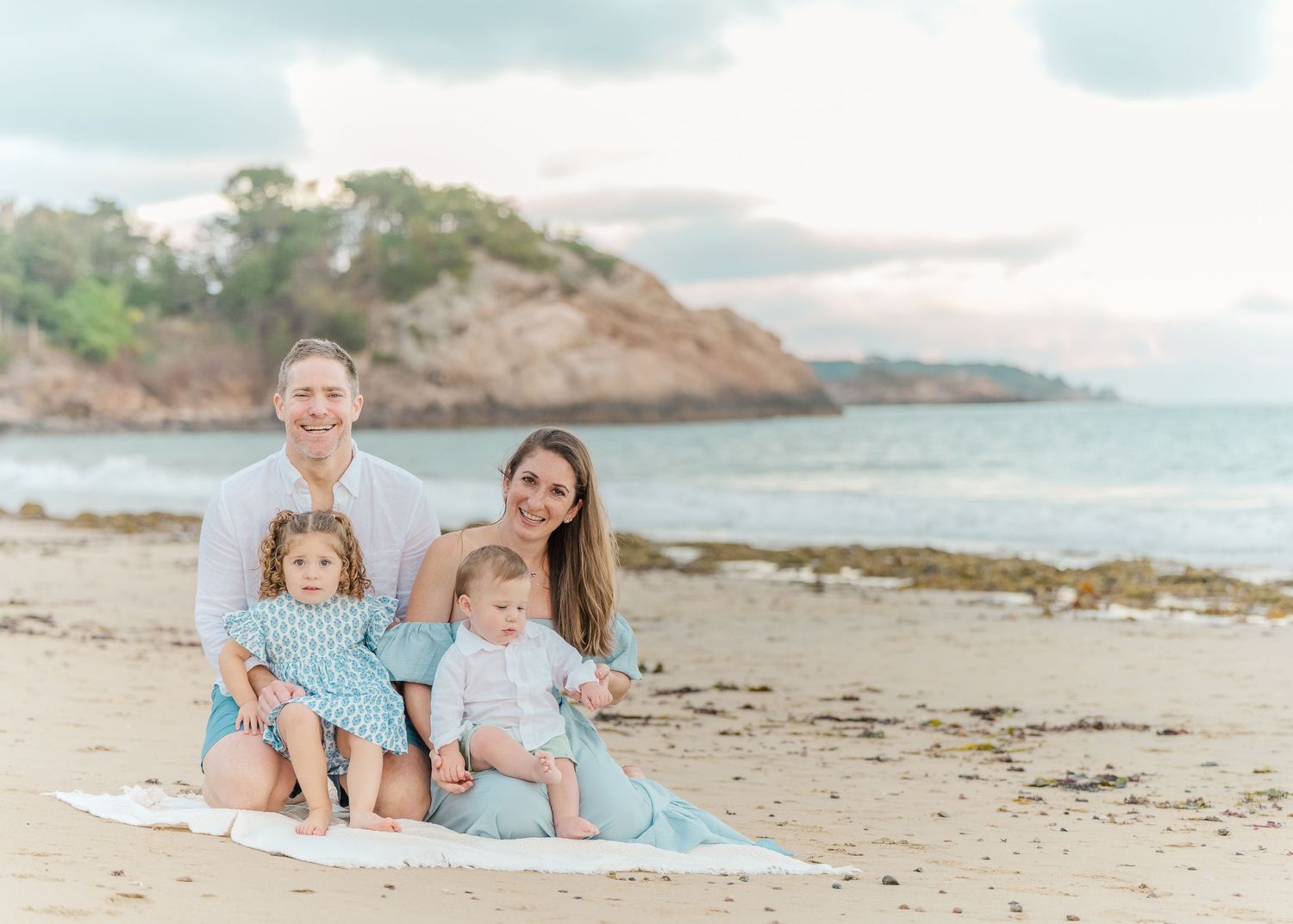 Family sitting together on blanket at scenic coastal beach with rocky cliffs and cloudy sky in background.